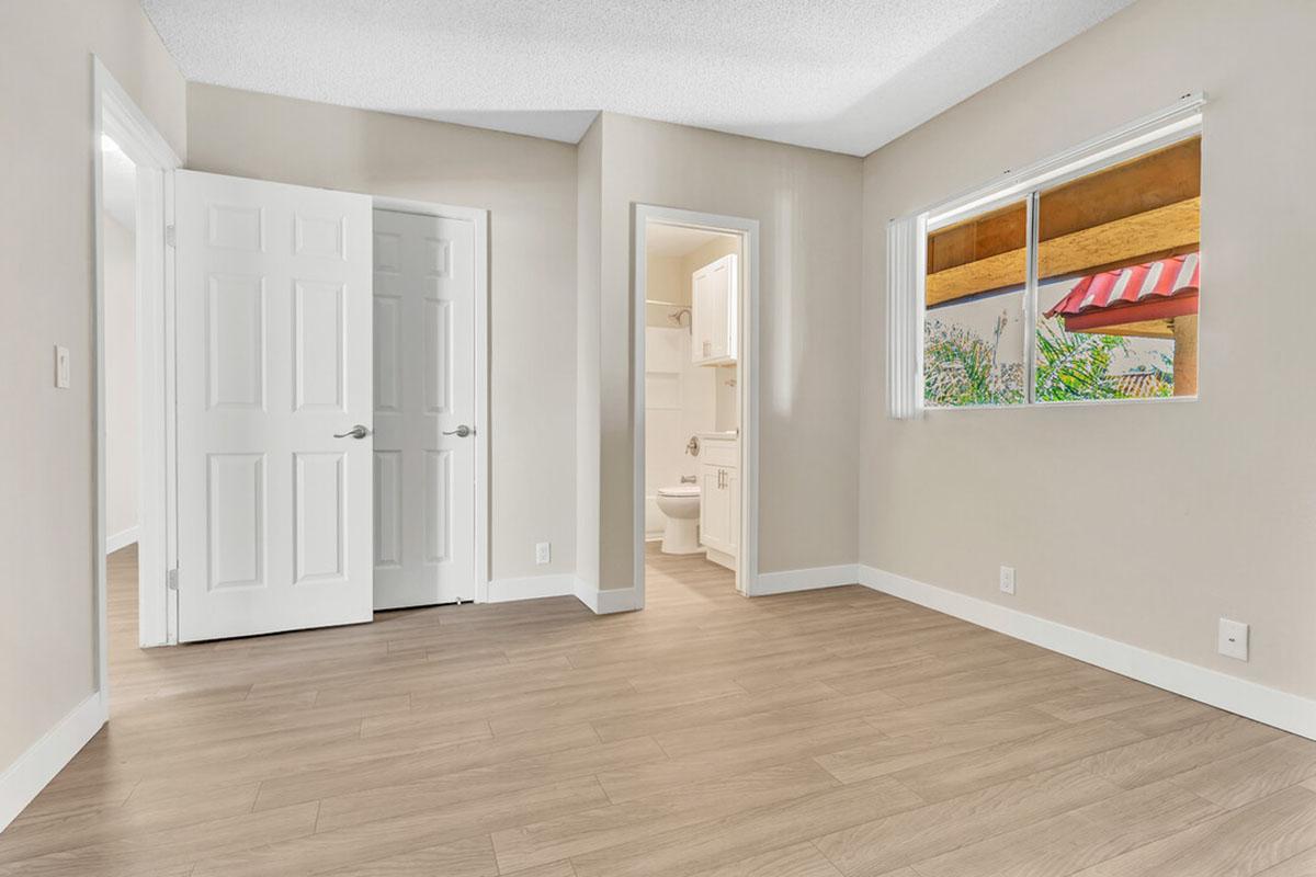 A well-lit, empty room with light-colored walls and wooden flooring. There are two open doors: one leading to a bathroom and the other to another space. A window with a view of greenery is also visible, adding to the bright atmosphere. The room has a clean, modern appearance.