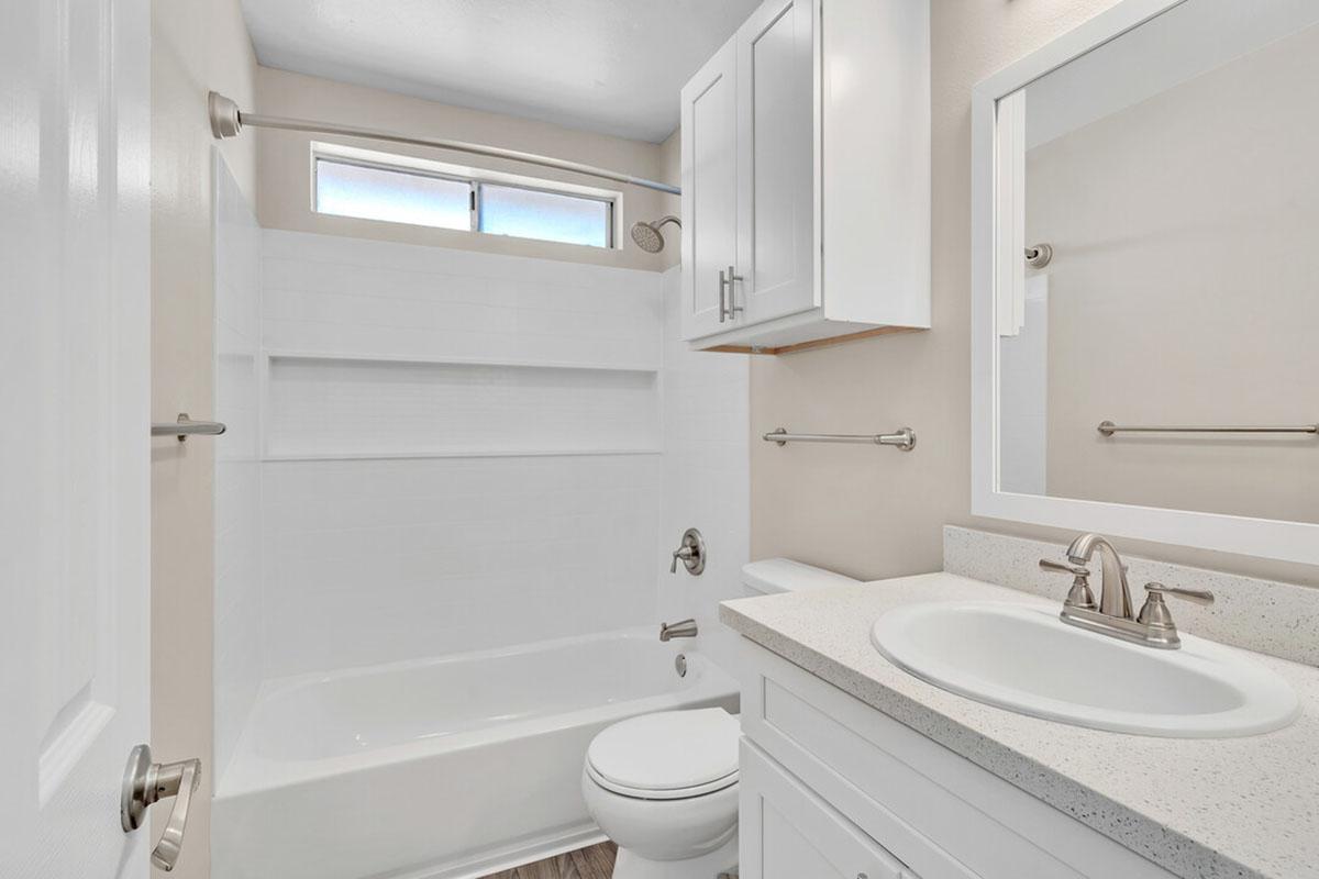 A clean and modern bathroom featuring a white bathtub and shower combination, a white vanity with a sink, a large mirror, and natural light coming through a window. The walls are painted in a light color, and the floor has a wood-like appearance.
