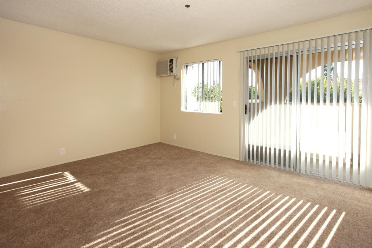 A spacious, empty room with beige walls and carpet. It features a window with vertical blinds allowing natural light to enter, casting shadows on the floor. An air conditioning unit is mounted on the wall, and large sliding glass doors lead to a balcony. The overall atmosphere is bright and inviting.