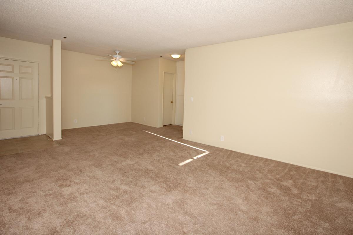 A spacious, unfurnished living room featuring beige walls and light brown carpet. There is a ceiling fan and a doorway leading to another room, with natural light entering through a possible window. The room appears clean and ready for furniture placement.