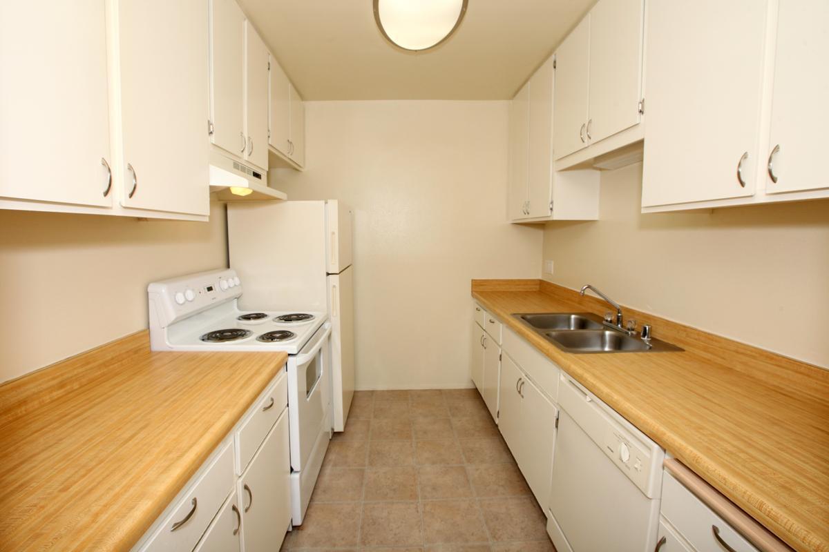 A small, modern kitchen featuring white cabinetry, a white stove and refrigerator, and a double sink. The countertops are light wood, and the floor has neutral tiles. The space is well-lit with natural light, creating a clean and inviting atmosphere.