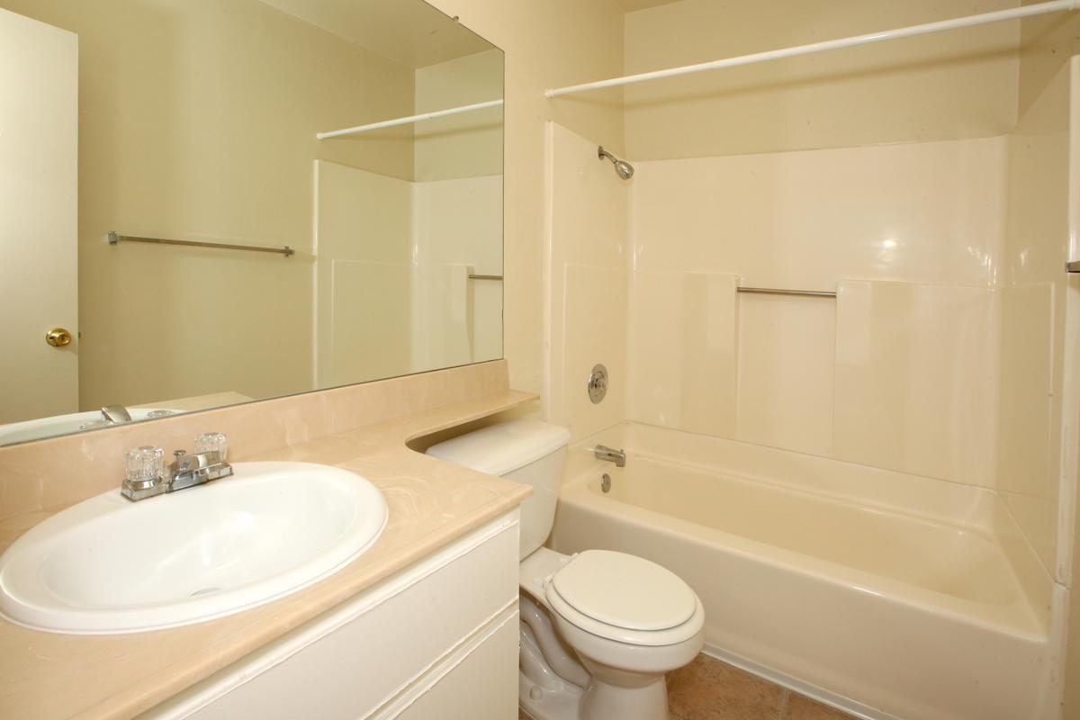 A clean, well-lit bathroom featuring a bathtub with a shower, a sink with a countertop, a mirror above the sink, and a toilet. The walls are painted a light color, and the floor has neutral tiles. A towel bar is mounted on the wall, and there is a door leading to another room.