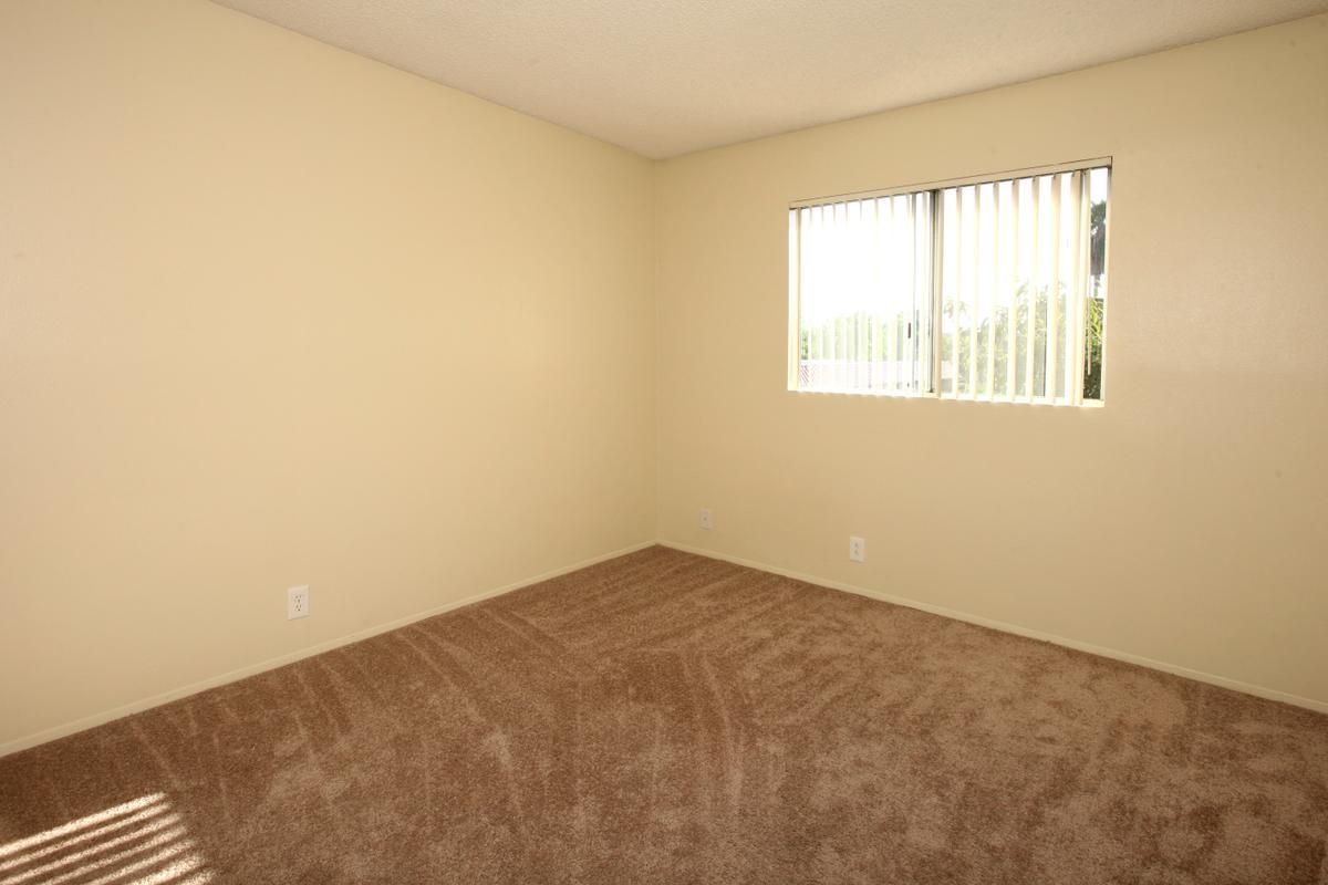 A vacant room with beige walls and light brown carpet. There is a window with vertical blinds on one wall, allowing natural light to enter the space. The room appears empty and unfurnished, creating a neutral and spacious atmosphere.