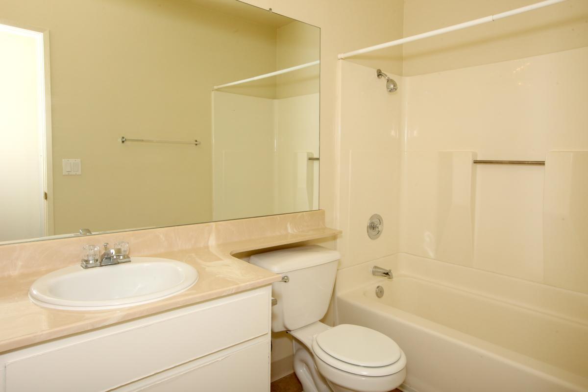 A clean, simple bathroom featuring a white bathtub with a shower, a toilet, and a sink with a beige countertop. The space is well-lit, with a large mirror above the sink and a towel bar mounted on the wall. The walls are a light, neutral color, creating a bright and airy feel.