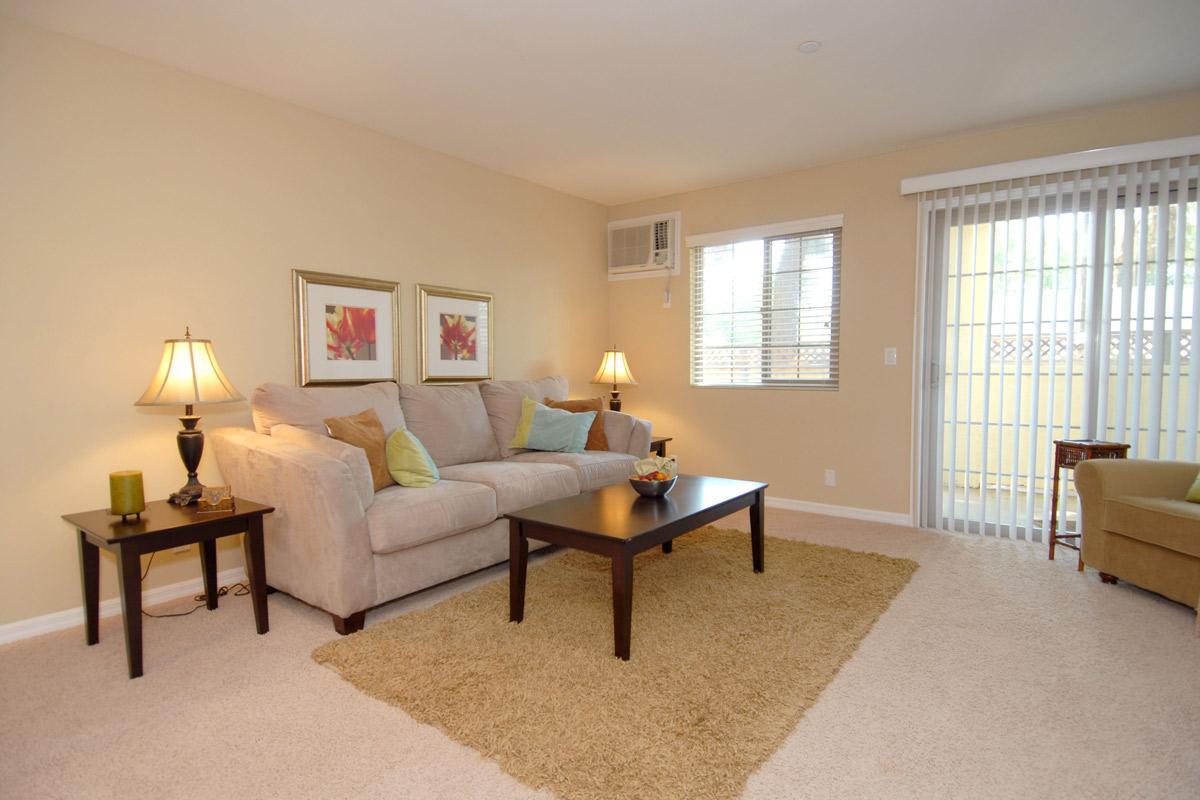 A cozy living room featuring a light beige sofa with colorful cushions, two lamps on side tables, a dark wood coffee table, and a small decorative bowl. There's a soft area rug on the beige carpet, a small green chair in the corner, and sliding glass doors with blinds leading to an outdoor area.