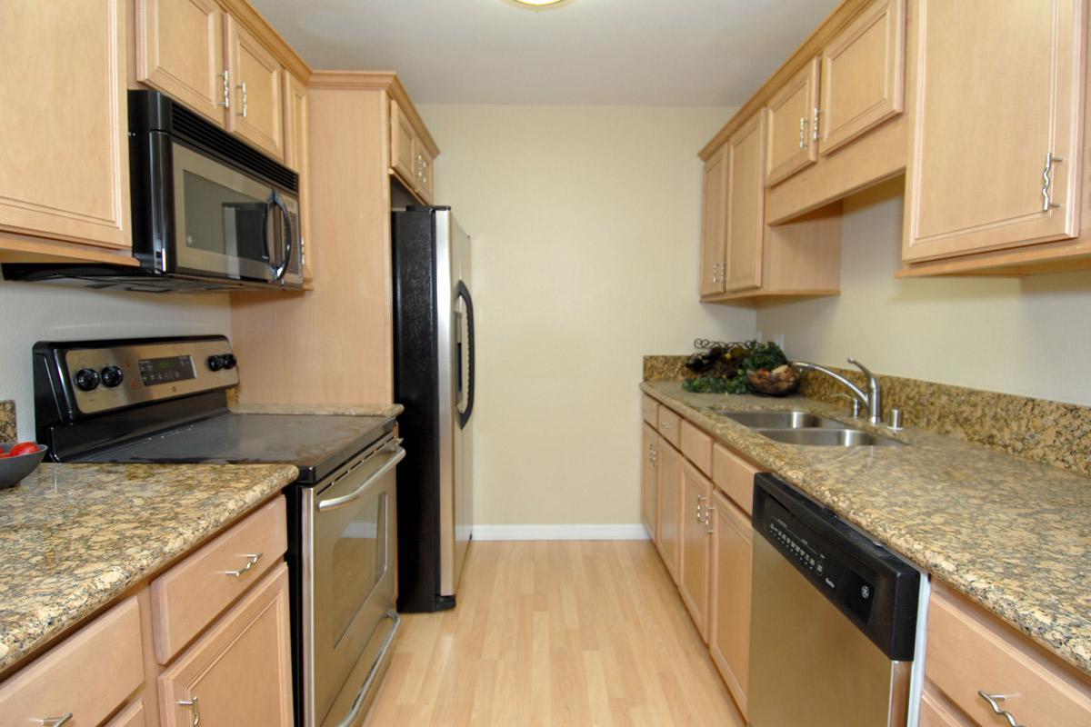 A modern kitchen featuring light wood cabinetry, granite countertops, and stainless steel appliances. The layout includes an oven, microwave, refrigerator, sink, and dishwasher. The flooring is light-colored wood, contributing to a bright and inviting atmosphere.