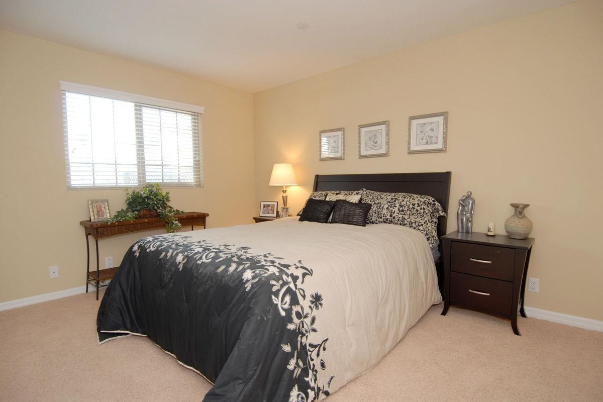 A cozy bedroom featuring a large bed with a decorative black and beige bedspread. The walls are painted a warm beige, and there are two framed pictures above the bed. A small wooden nightstand holds a lamp, and a decorative vase is placed nearby. Natural light enters through a window with closed blinds.