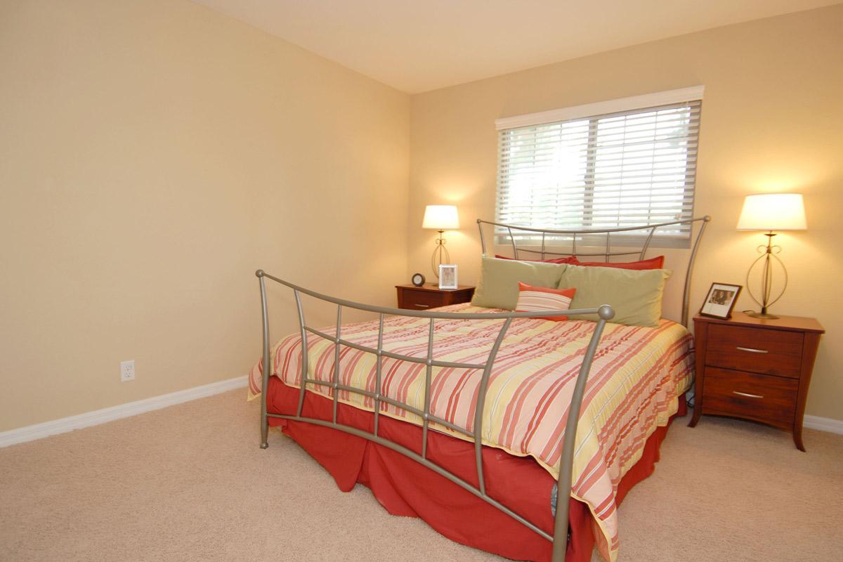 A cozy bedroom featuring a metal bed frame with a colorful striped comforter, two bedside lamps, and a wooden nightstand. The walls are painted a soft beige, and a window with blinds allows natural light to filter in, creating a warm and inviting atmosphere. A framed photo decorates the nightstand.