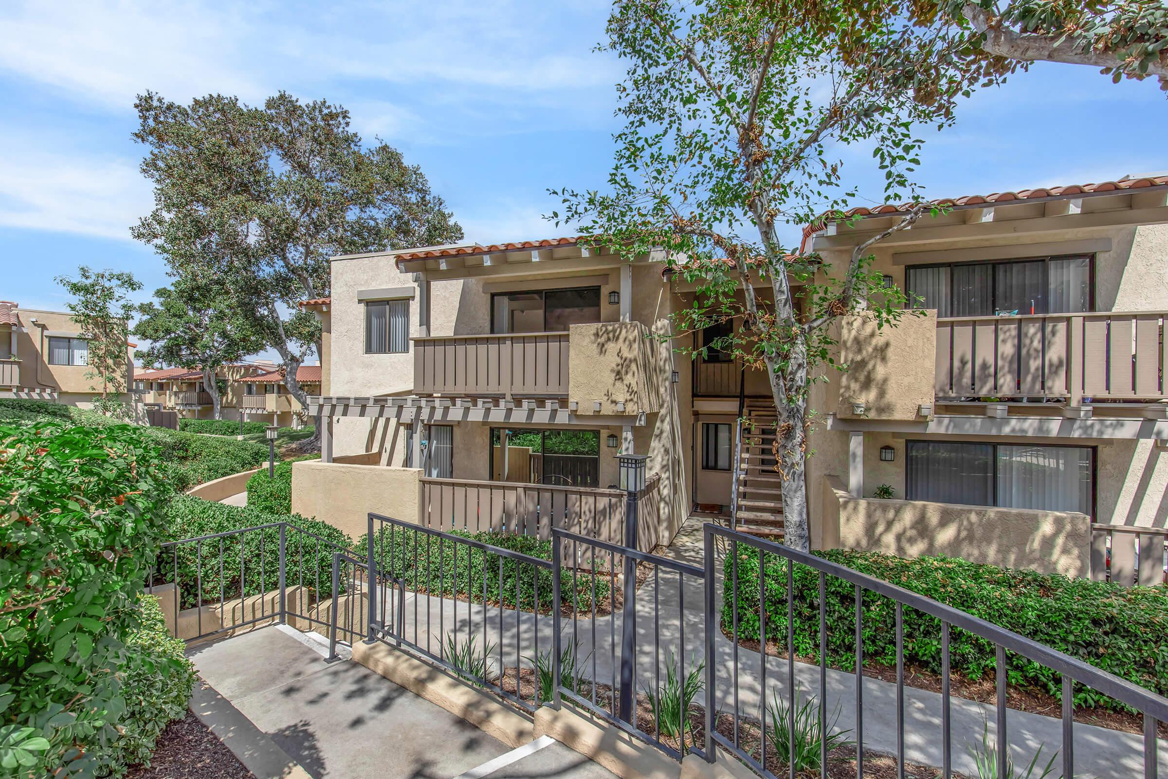 A view of a multi-unit apartment building surrounded by greenery. The image shows a path leading to the entrance, with balconies visible on the upper floors and trees in the background under a clear blue sky. The atmosphere conveys a peaceful residential environment.