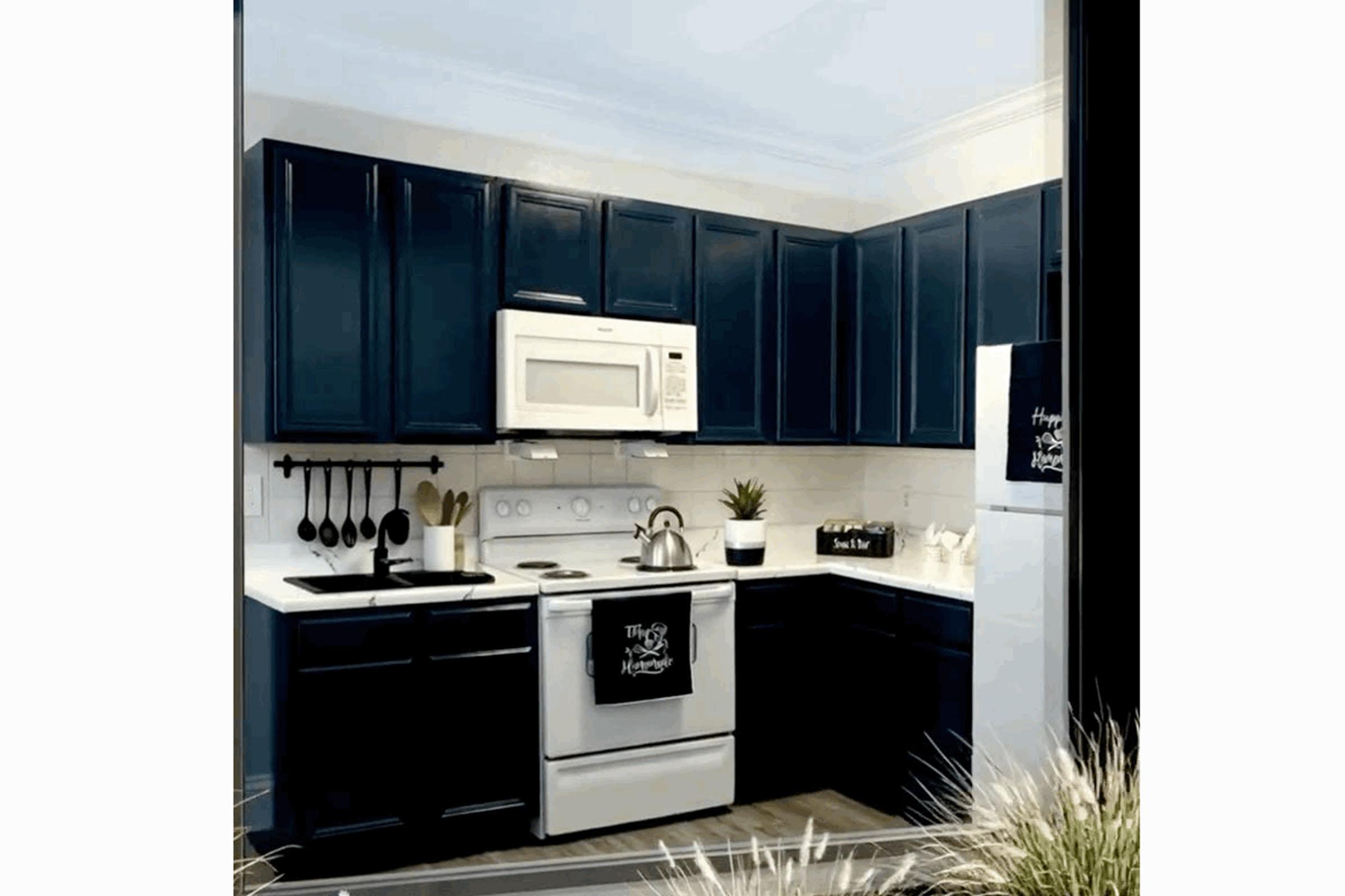 A modern kitchen featuring dark blue cabinets, a white countertop, a white microwave above the stove, and stainless steel appliances. The kitchen includes a black utensil holder, a plant, and decorative signs. The space is well-lit and organizes kitchen essentials neatly.
