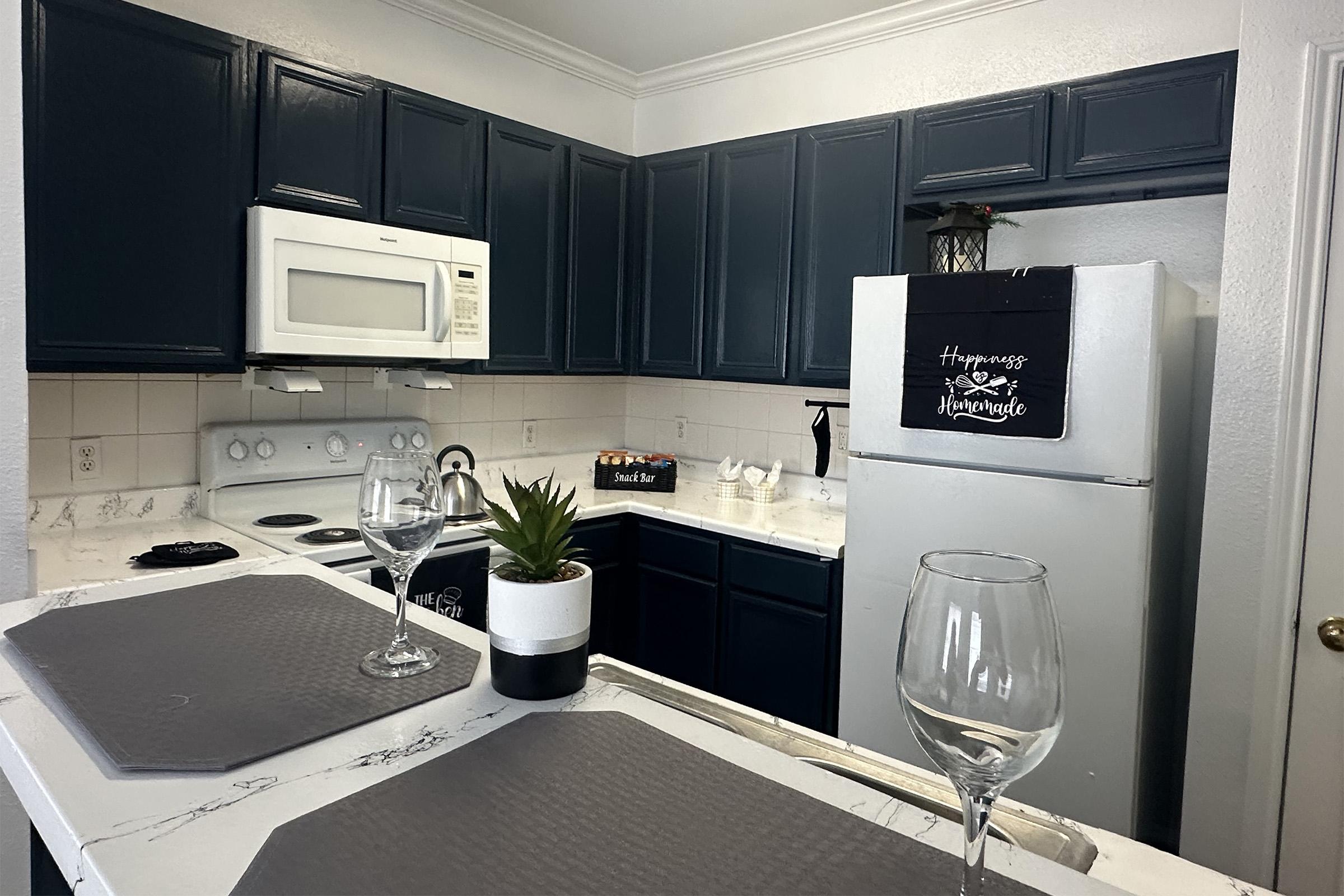 A modern kitchen featuring dark blue cabinets, a white countertop, and stainless steel appliances. A microwave and a white stove are visible. Two wine glasses and a small potted plant sit on the island, with a decorative sign on the refrigerator. The space is well-lit and organized, creating a welcoming atmosphere.