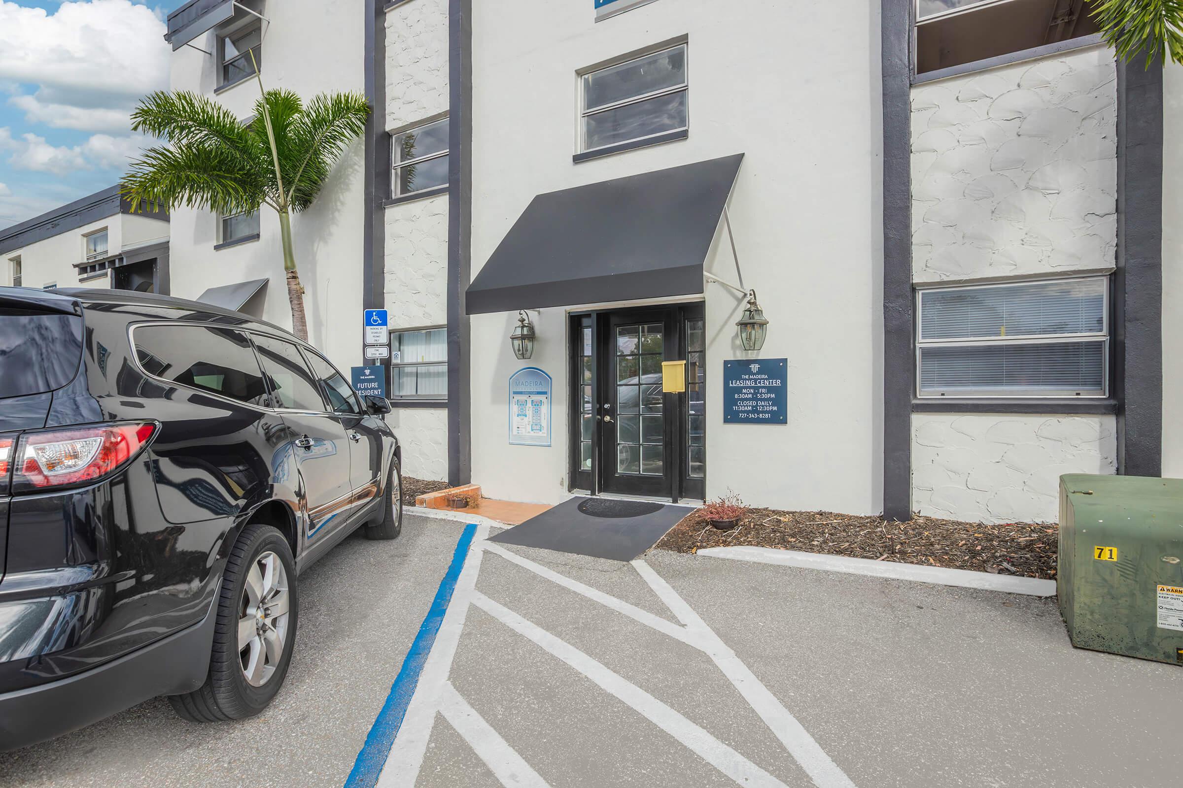 Exterior view of a building entrance featuring a dark awning, a door with a glass panel, and nearby signage. A black SUV is parked next to the entrance, with visible markings on the ground for parking spaces. Palm trees are seen in the foreground against a partly cloudy sky.