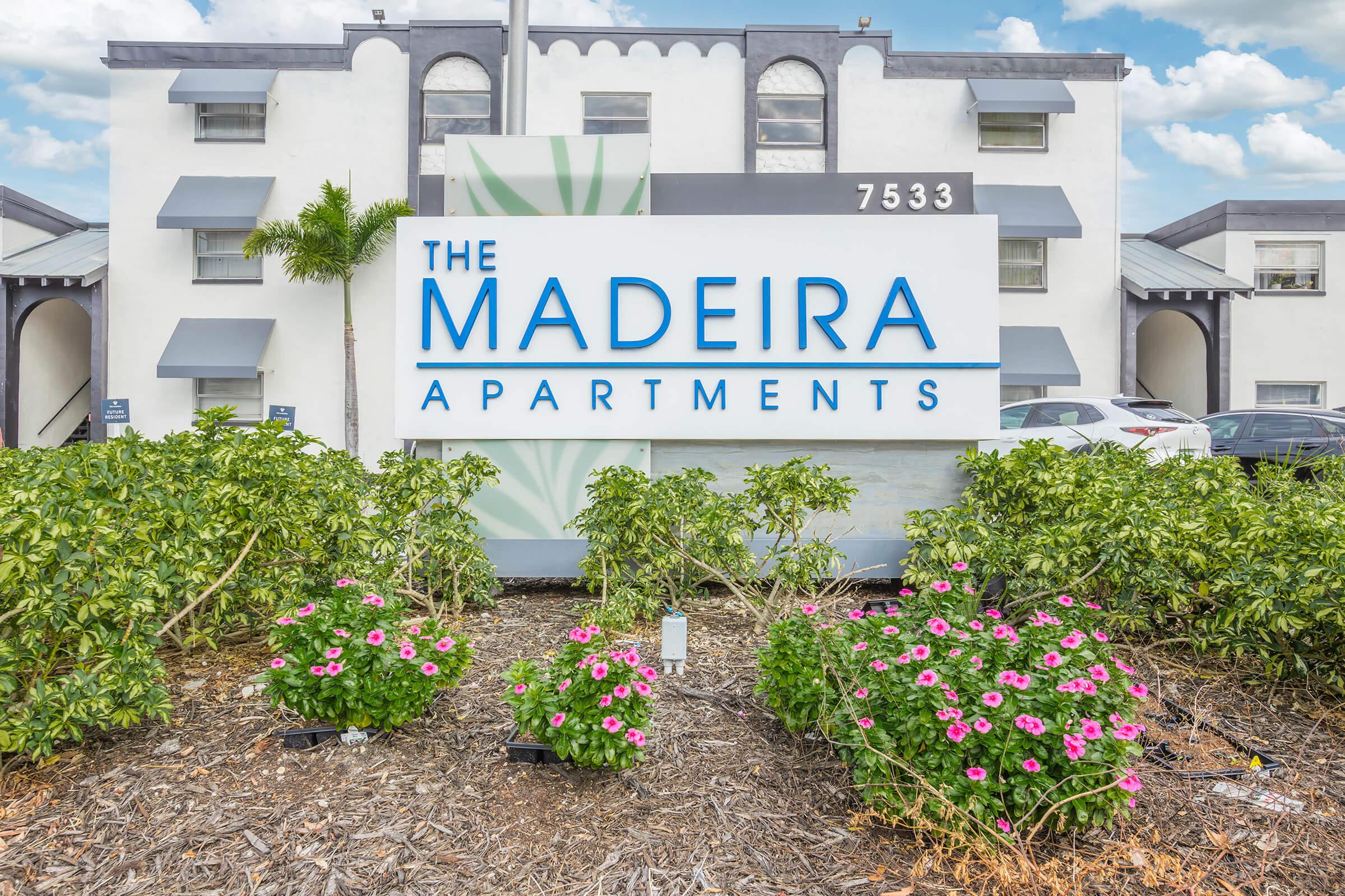 Sign for The Madeira Apartments, featuring large blue lettering against a light background. The sign is surrounded by green landscaping and colorful pink flowers. In the background, a multi-story apartment building is visible under a partly cloudy sky.