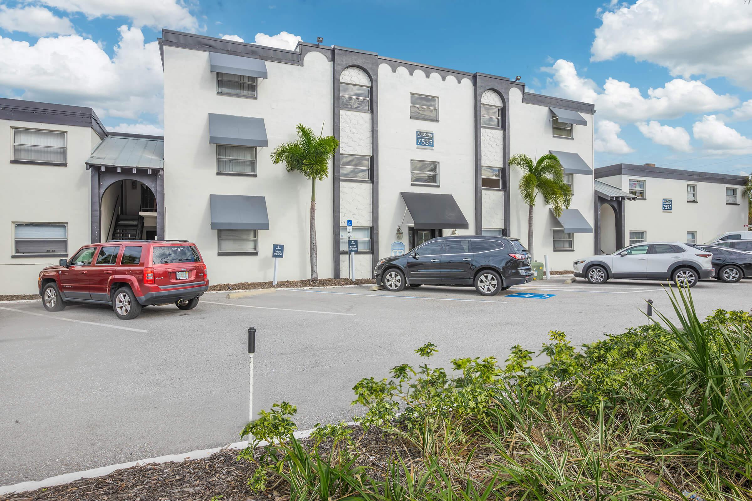 A two-story white apartment building with several windows and gray awnings. In the foreground, there's a parking lot with a variety of cars, including a red SUV and a black SUV. The landscape features green shrubs and palm trees, under a partly cloudy blue sky.