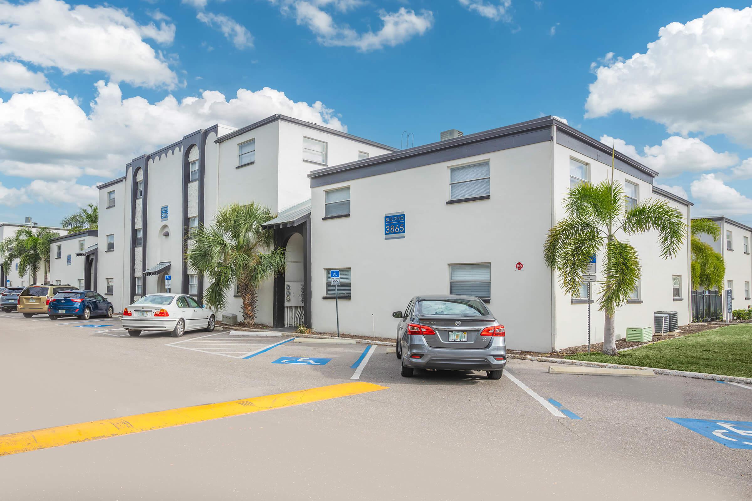 A view of a white multi-unit residential building with a palm tree in the foreground. The building features multiple windows and a sign. Several cars are parked in designated spaces, including a grey sedan and other vehicles. The sky is partly cloudy, adding to the bright and welcoming atmosphere.