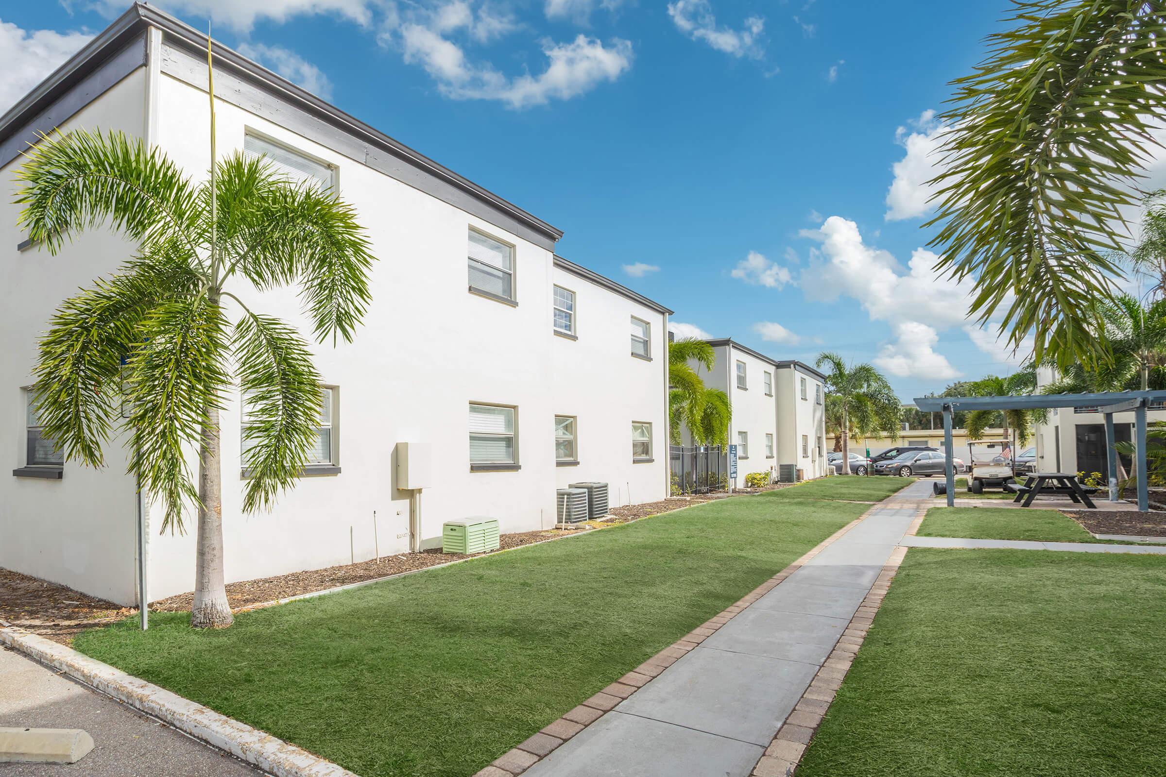 View of a well-maintained apartment complex featuring two-story white buildings surrounded by green grass and palm trees. A paved walkway runs through the lawn, leading to picnic areas and additional greenery, under a clear blue sky with scattered clouds.