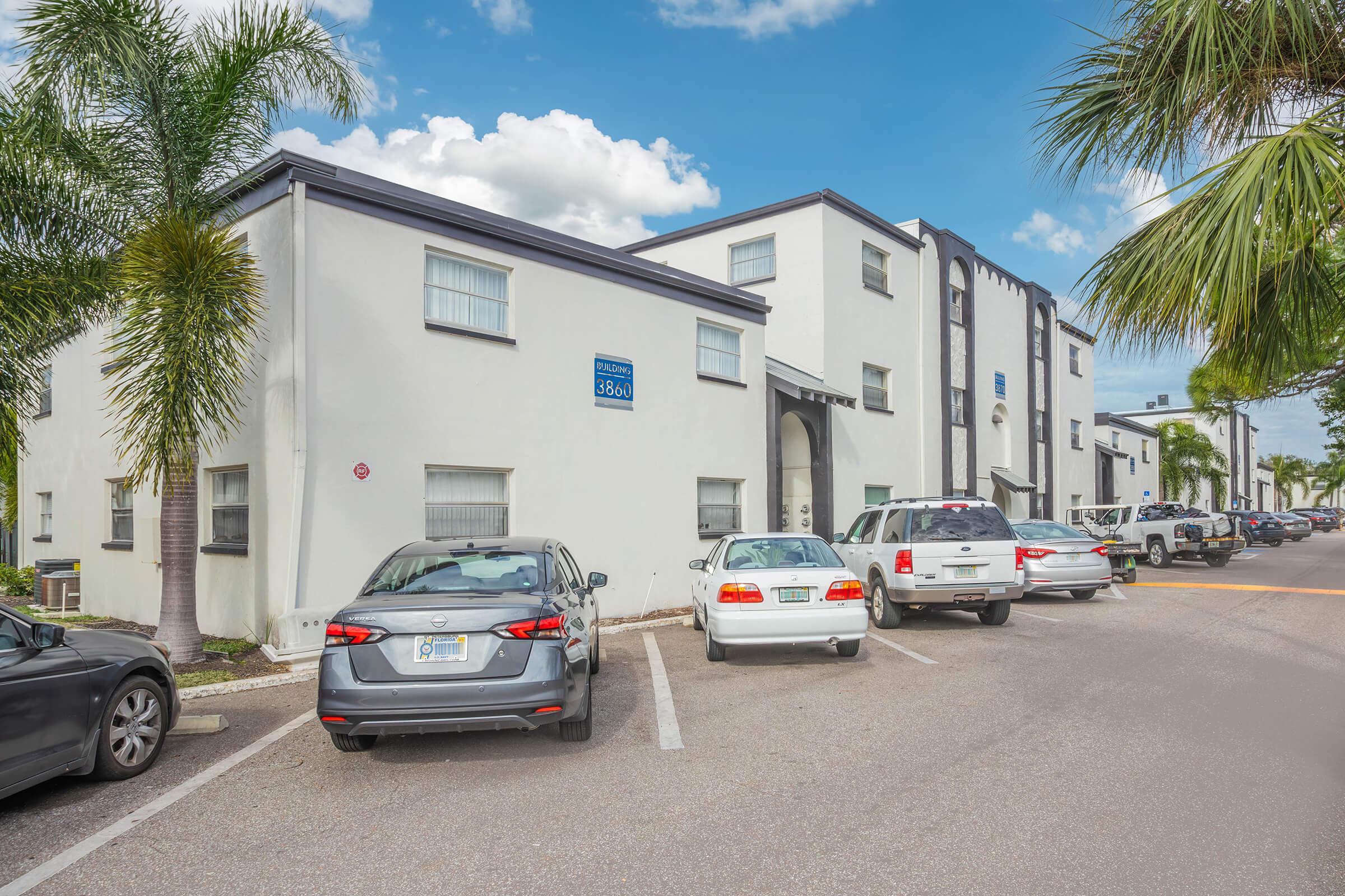 Exterior view of a white, two-story apartment building with several parked cars in the foreground. Palm trees line the parking area, and the sky is partly cloudy. A sign on the building indicates its address. The scene portrays a residential setting in a warm climate.