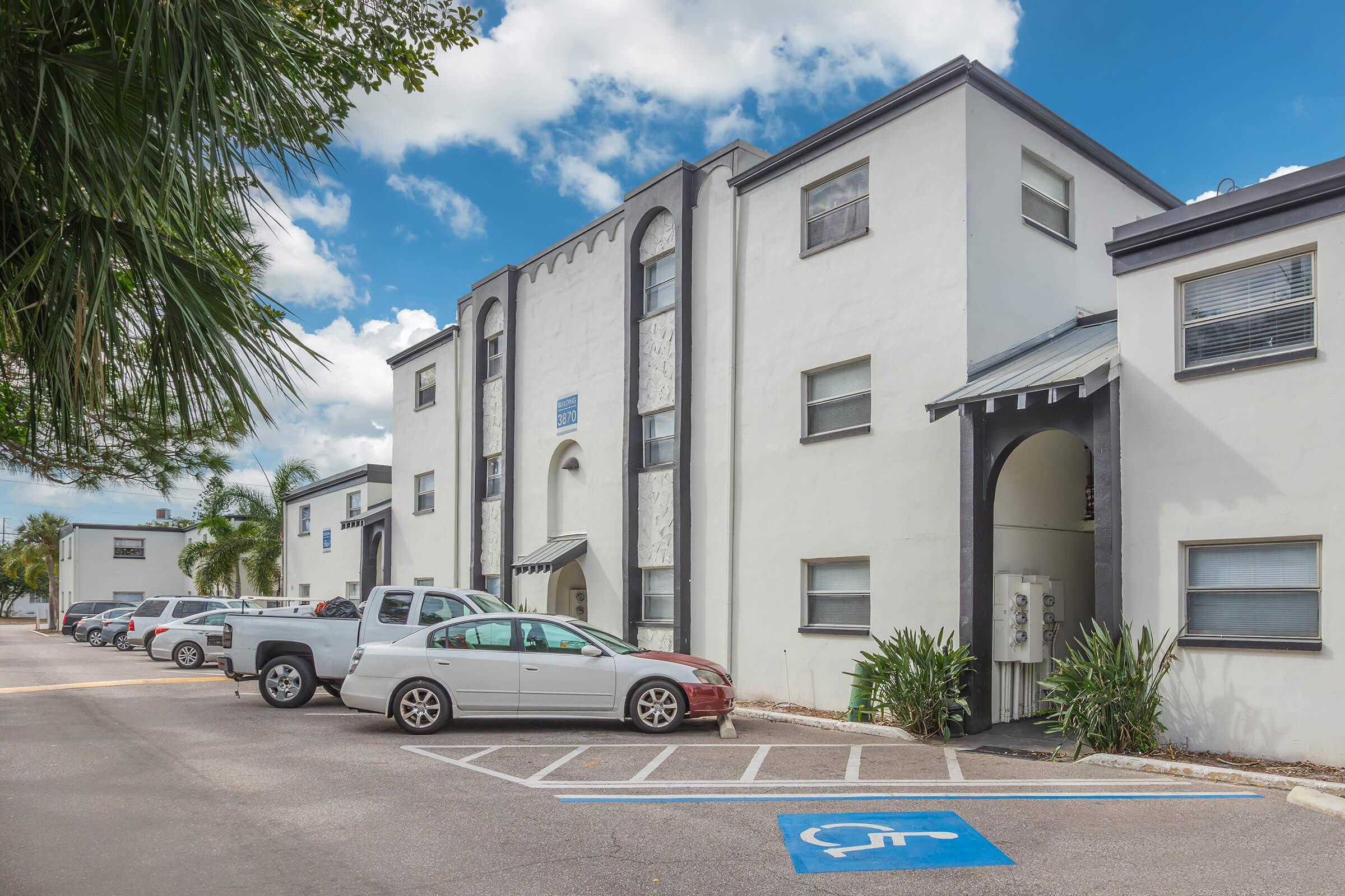 A view of a two-story white apartment building with a modern architectural style. The parking lot features several vehicles, including a red car and a white SUV. Lush greenery surrounds the area, and a blue sky with clouds is visible above.