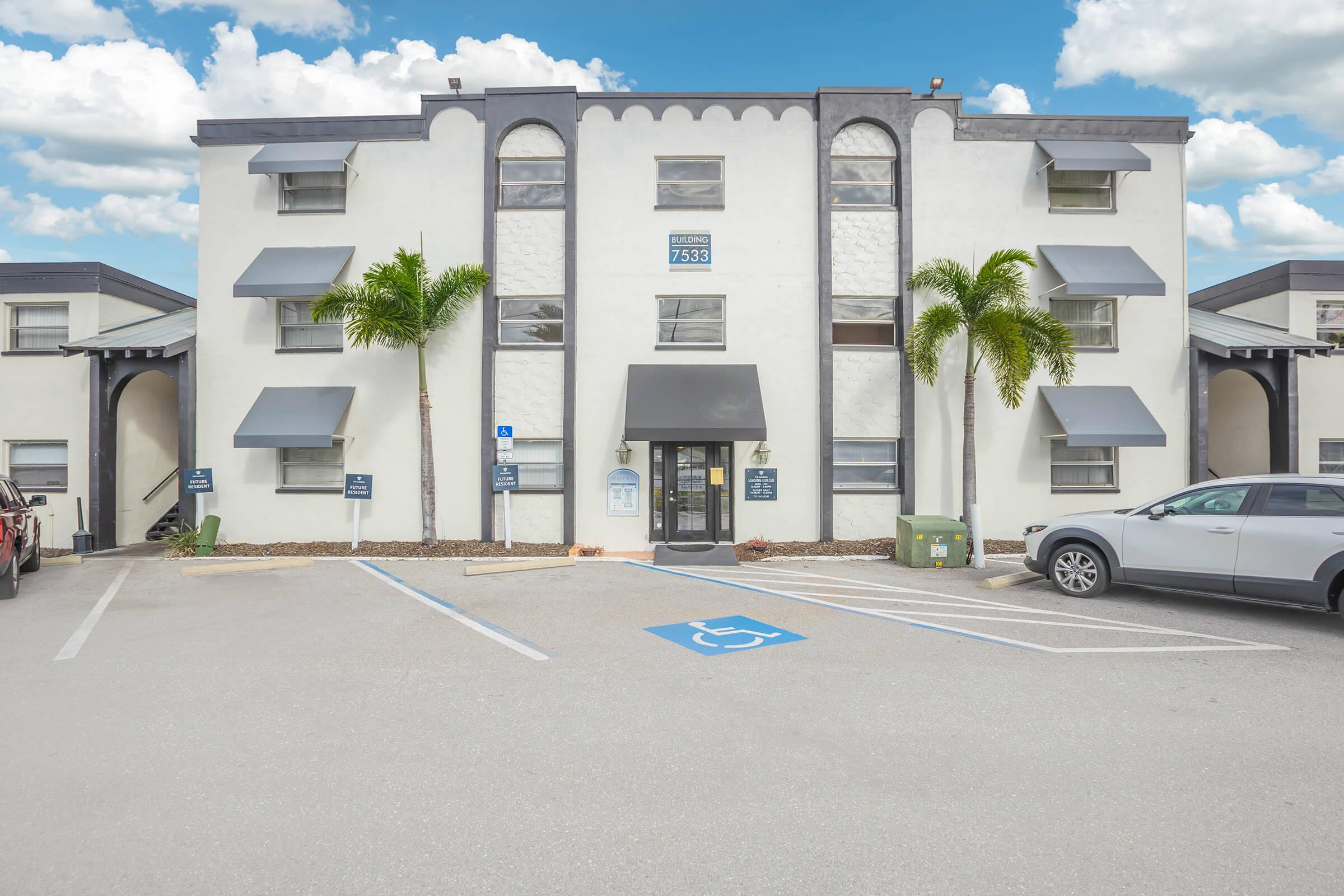 Three-story white building with a black awning and large windows, flanked by palm trees. The entrance is centrally located, with several parking spaces in front, including a designated handicapped space. The sky is partly cloudy.