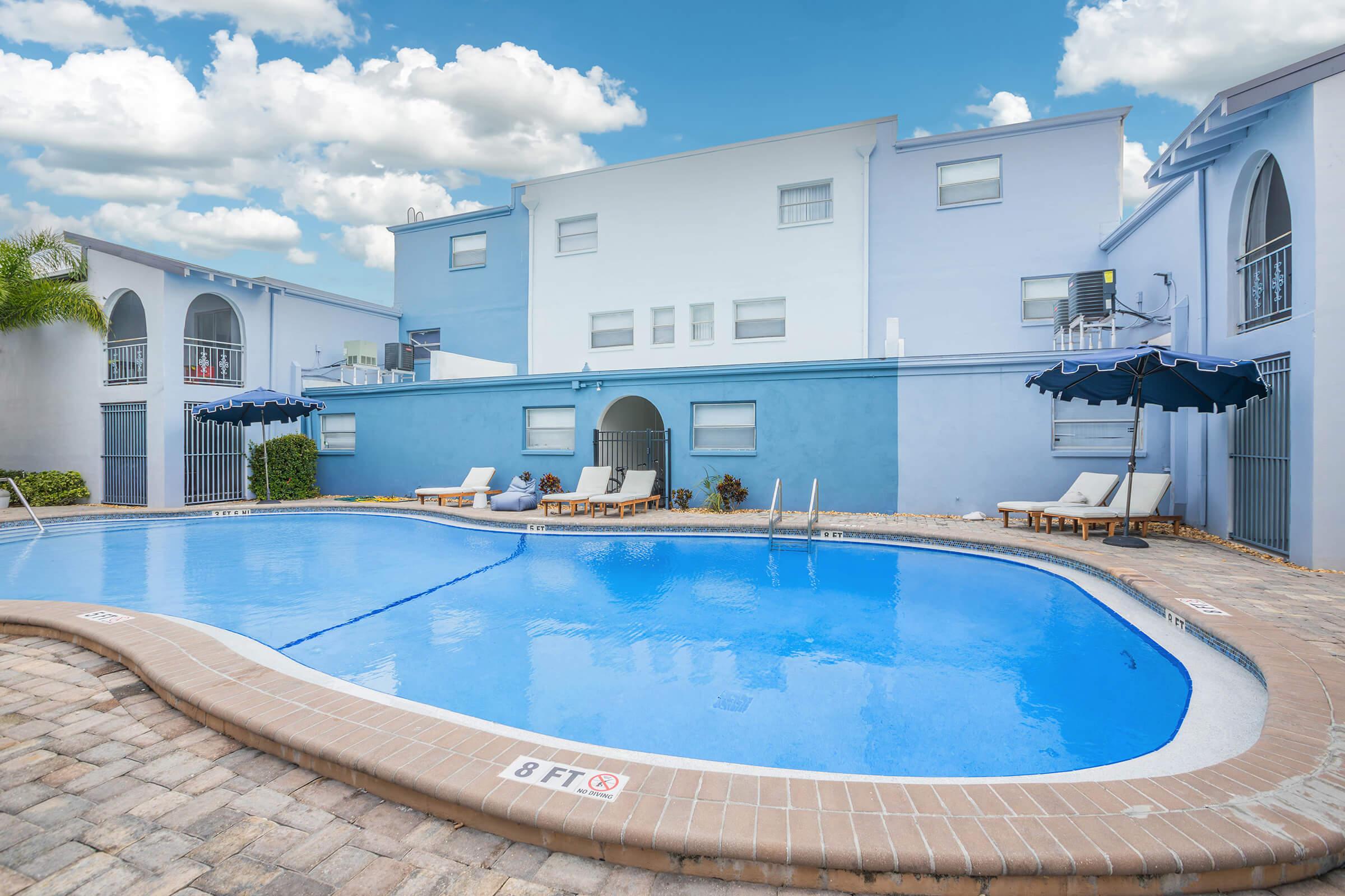 A bright blue pool surrounded by lounge chairs and umbrellas, with a light blue building in the background. The scene is set under a partly cloudy sky, creating a relaxing atmosphere. The pool area features a decorative stone border and clear water, inviting for a swim.