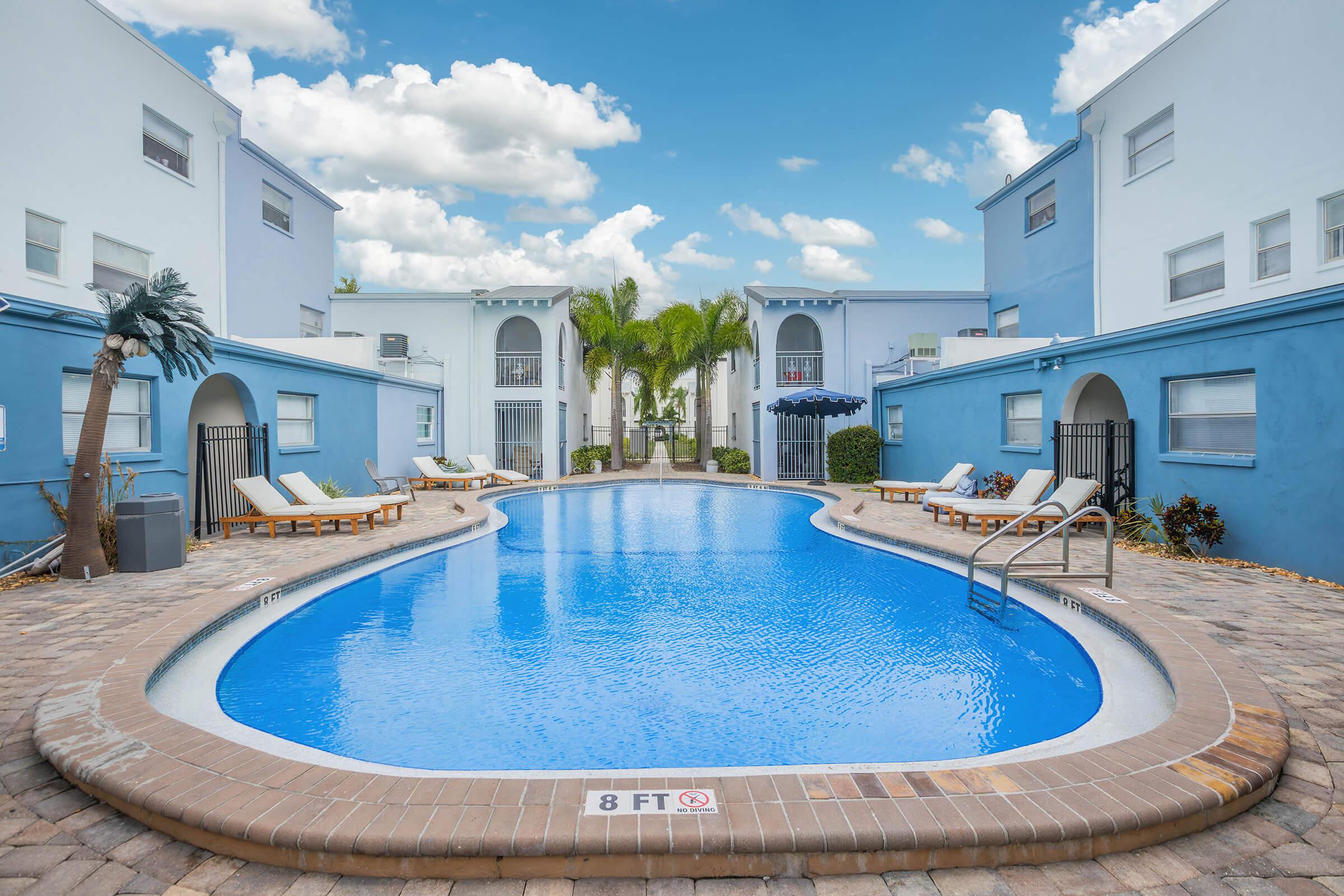 View of a blue swimming pool surrounded by lounge chairs and palm trees, located in a residential area. The pool has clear water, and a "8 FT" depth marker is visible. In the background, there are white and blue buildings under a partly cloudy sky, creating a serene, inviting atmosphere.