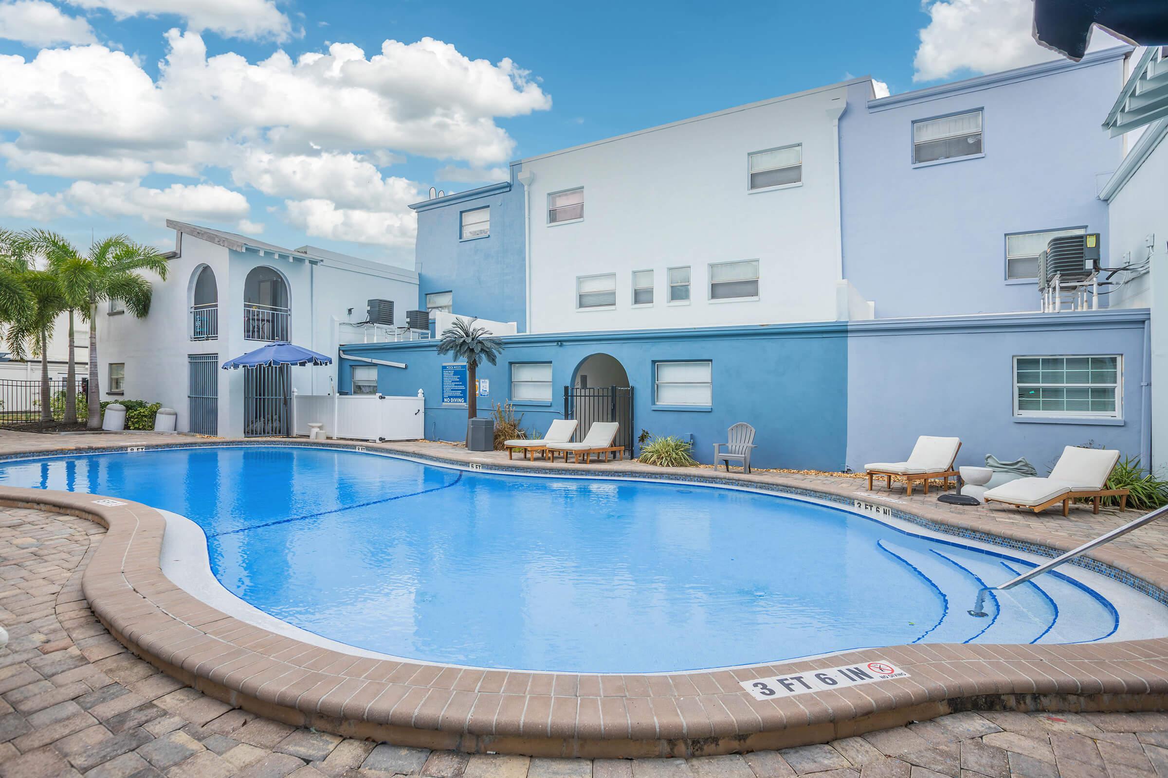A serene outdoor swimming pool area with a light blue building in the background. The pool features a gradual slope, sun loungers beside it, and an umbrella for shade. Palm trees add a tropical touch against a partly cloudy sky. The scene is inviting and perfect for relaxation.