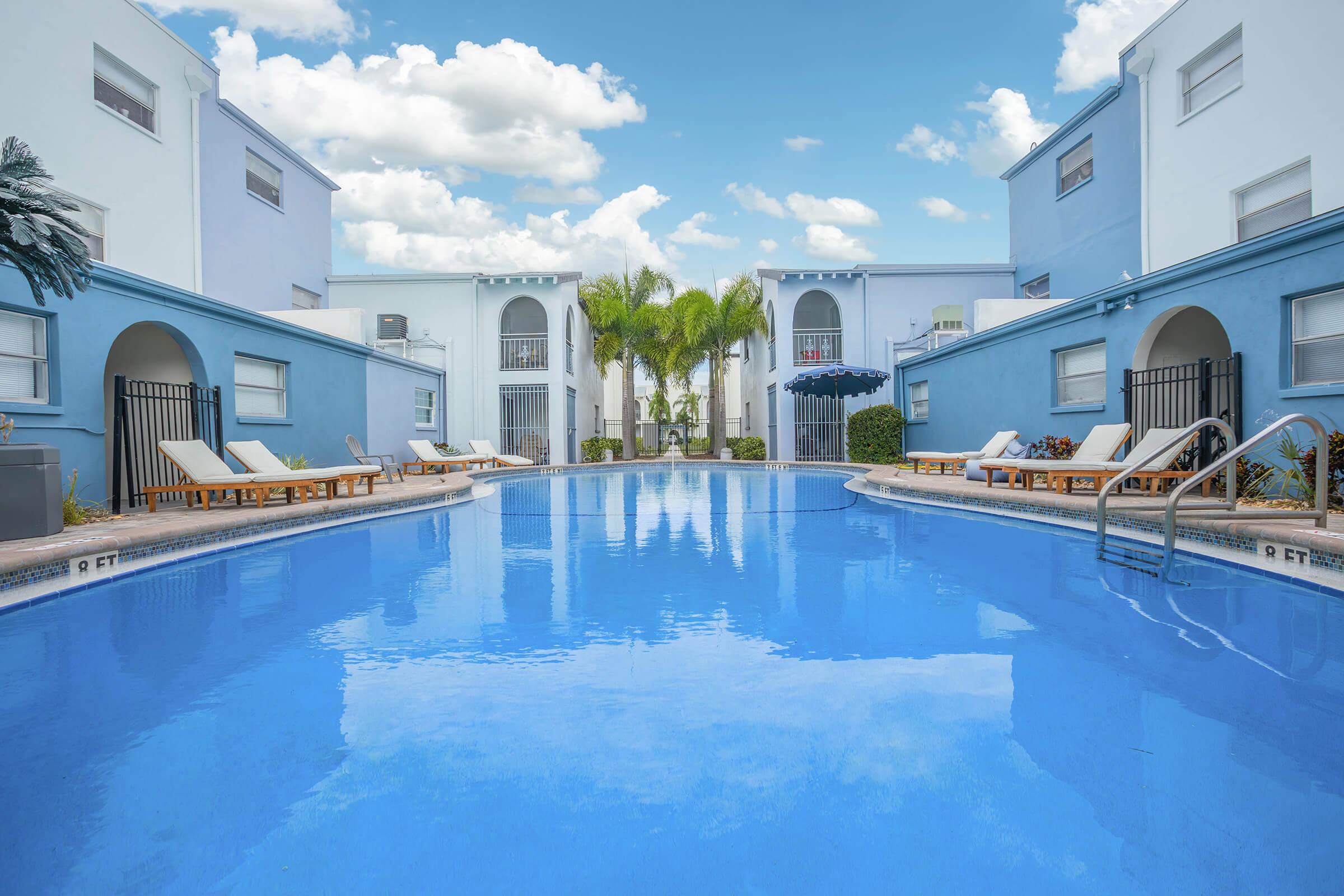 A serene swimming pool surrounded by light blue buildings, lounge chairs, and palm trees under a partly cloudy sky. The pool reflects the blue of the sky, creating a tranquil and inviting atmosphere for relaxation and leisure.