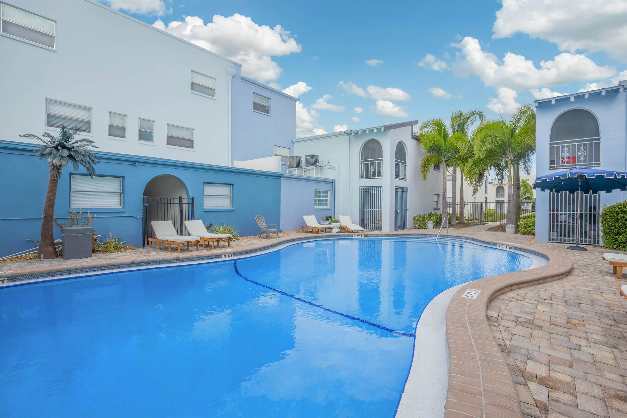A swimming pool surrounded by lounge chairs and palm trees, with blue buildings and palm trees in the background. The sky is bright with fluffy clouds, creating a relaxing atmosphere.