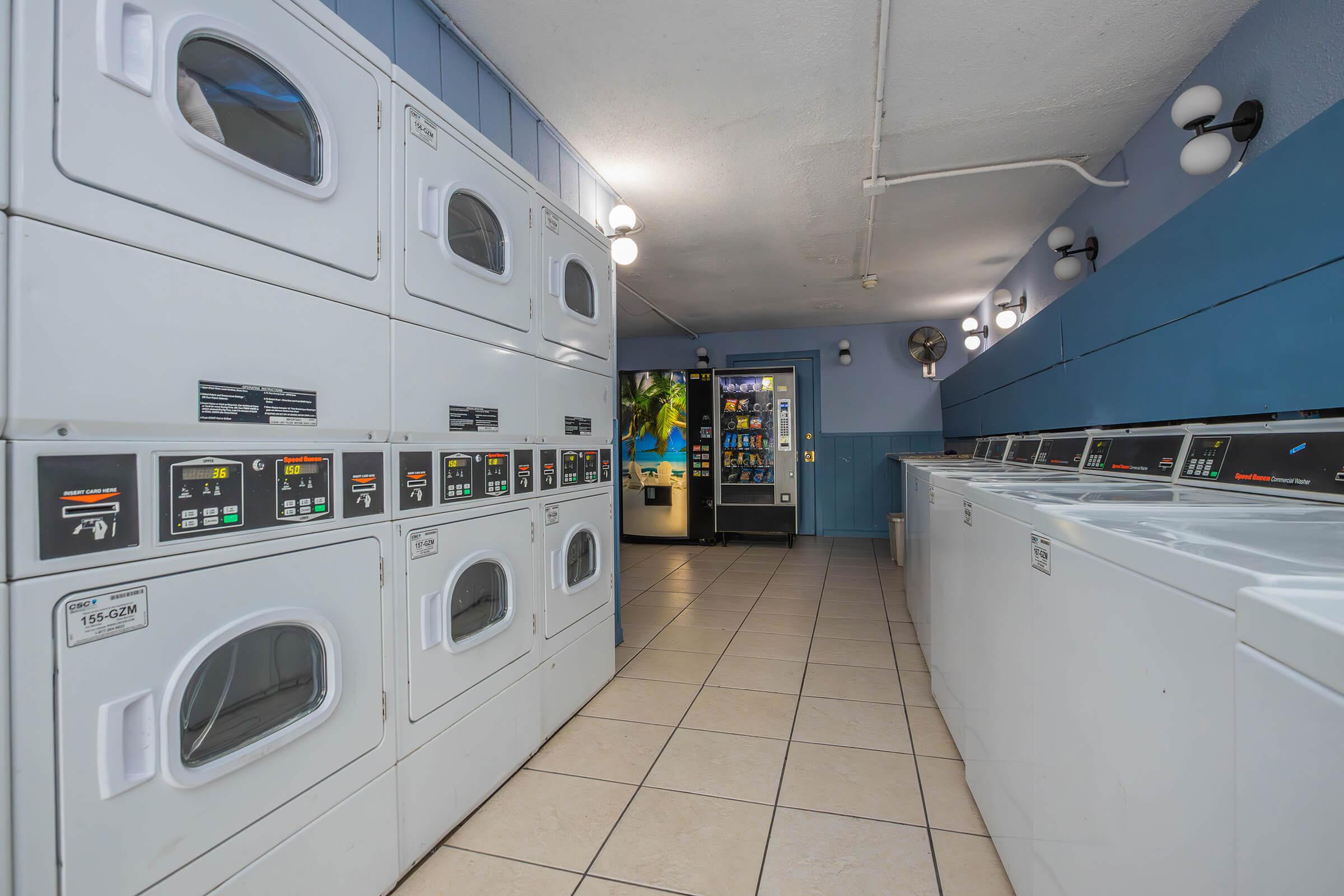 A clean laundromat interior featuring rows of stacked washing machines and dryers. The tiled floor and blue walls create a bright atmosphere. In the background, a vending machine can be seen, providing snacks and drinks for customers. Soft lighting fixtures enhance the welcoming environment.