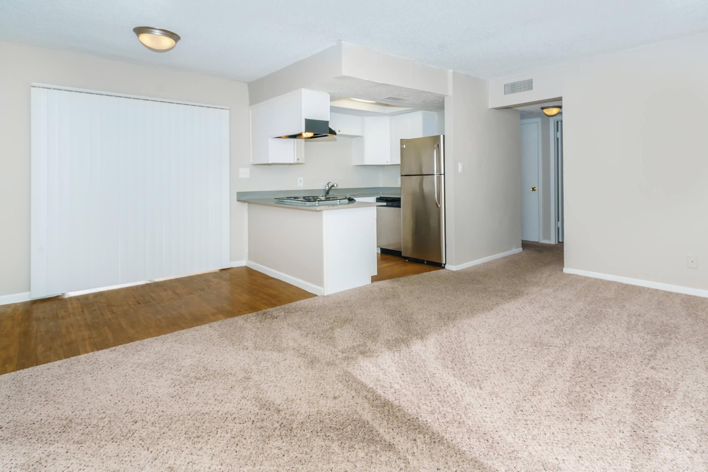 Spacious living area with light-colored walls and beige carpet. Features a kitchen with white cabinets, a stainless steel refrigerator, and a sliding glass door leading to a patio. The layout includes an open space suitable for dining and living, enhancing the airy and bright atmosphere.