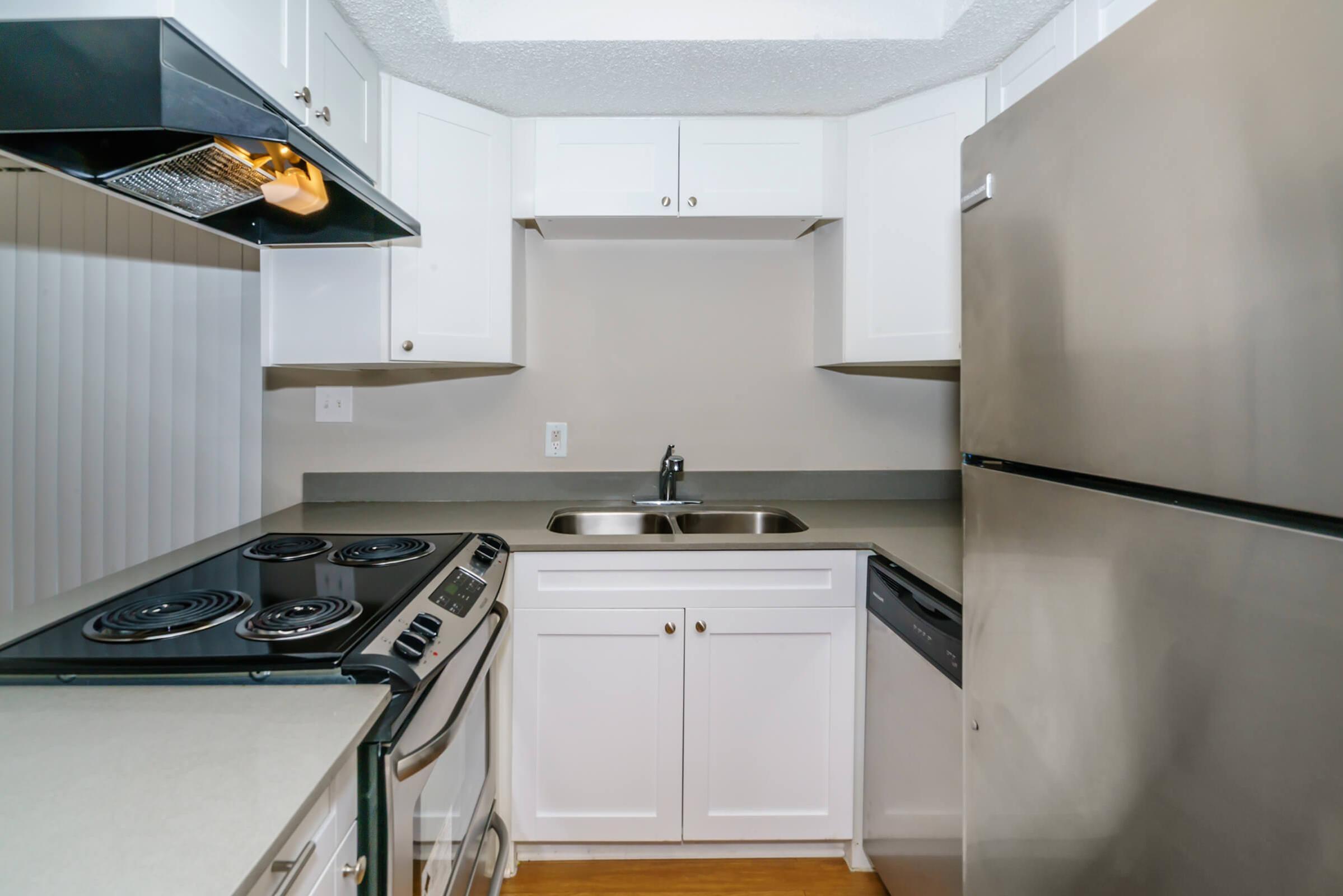 A modern kitchen featuring white cabinetry, a stainless steel refrigerator, an oven with a stove top, and a double-basin sink. The countertop is light-colored, and the walls are painted in a neutral shade, creating a clean and bright appearance.