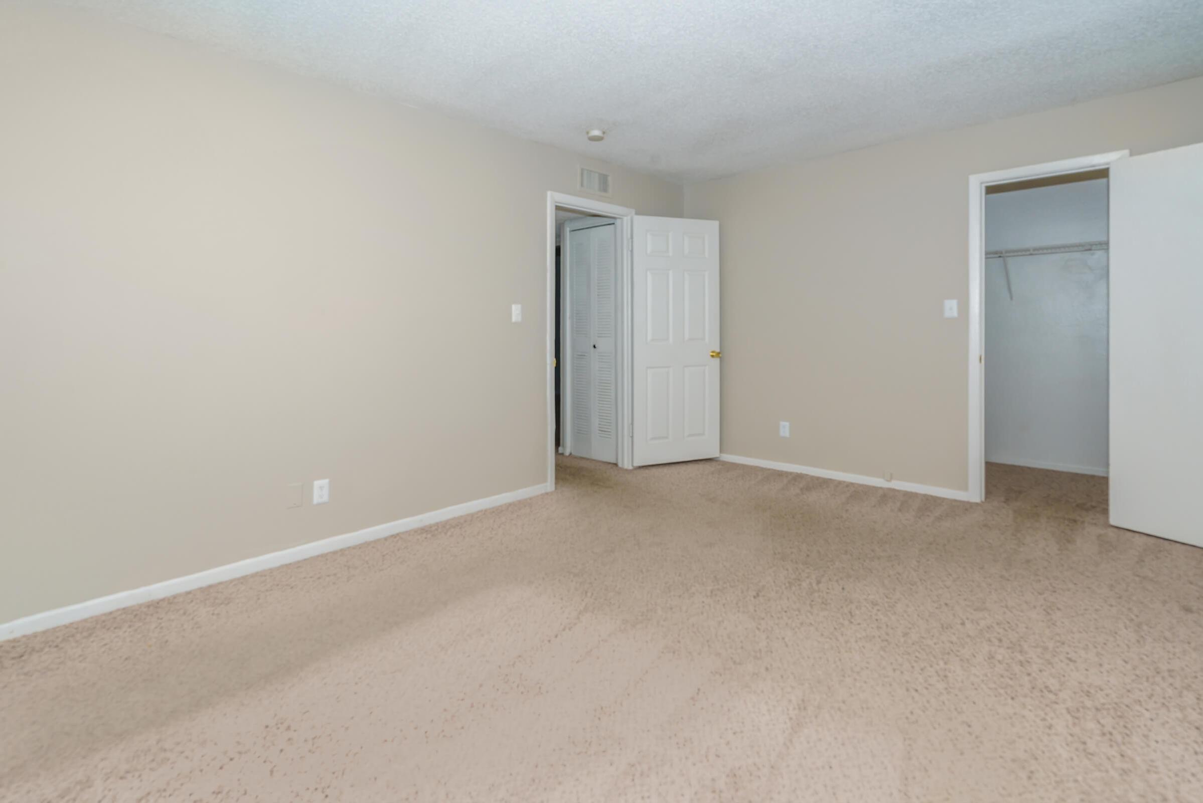 Empty room featuring beige walls and carpet, with a white closet door visible on the right. A second door leads to another area, and the ceiling has a textured finish. Natural light brightens the space, creating a spacious, neutral atmosphere.
