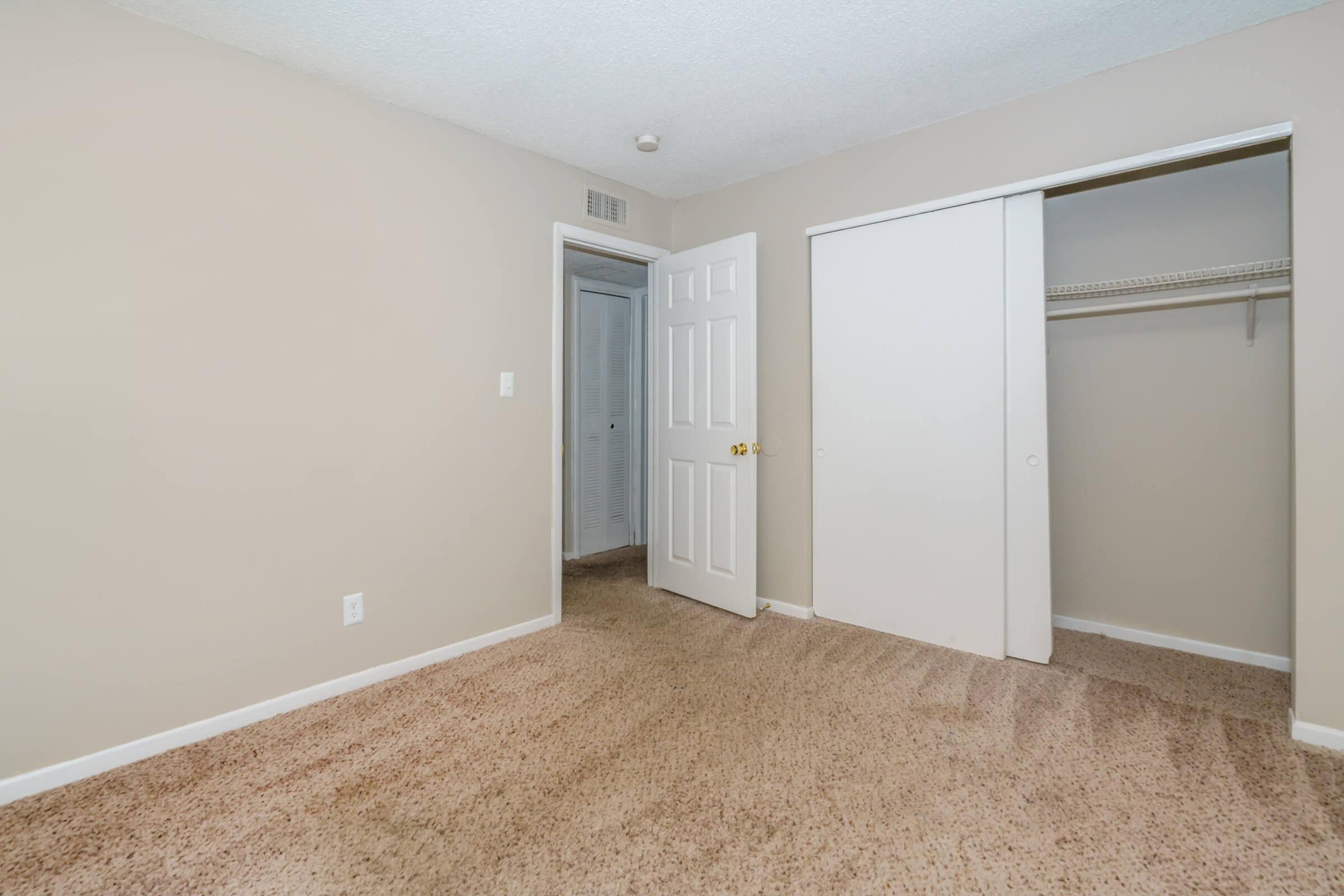 A simple, empty room with beige walls and carpeted flooring. There is a closet with sliding doors on the right and a doorway leading to another space in the background. Natural light illuminates the room, creating a bright and open atmosphere. Perfect for a bedroom or personal office.