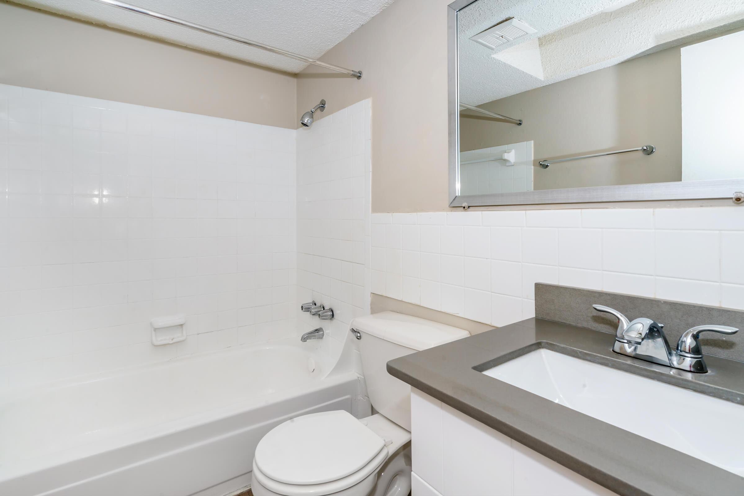 A clean, modern bathroom featuring a white bathtub, a toilet, and a sleek sink with a dark countertop. The walls are light-colored tiles, and there is a large mirror above the sink. The space appears well-lit and organized.