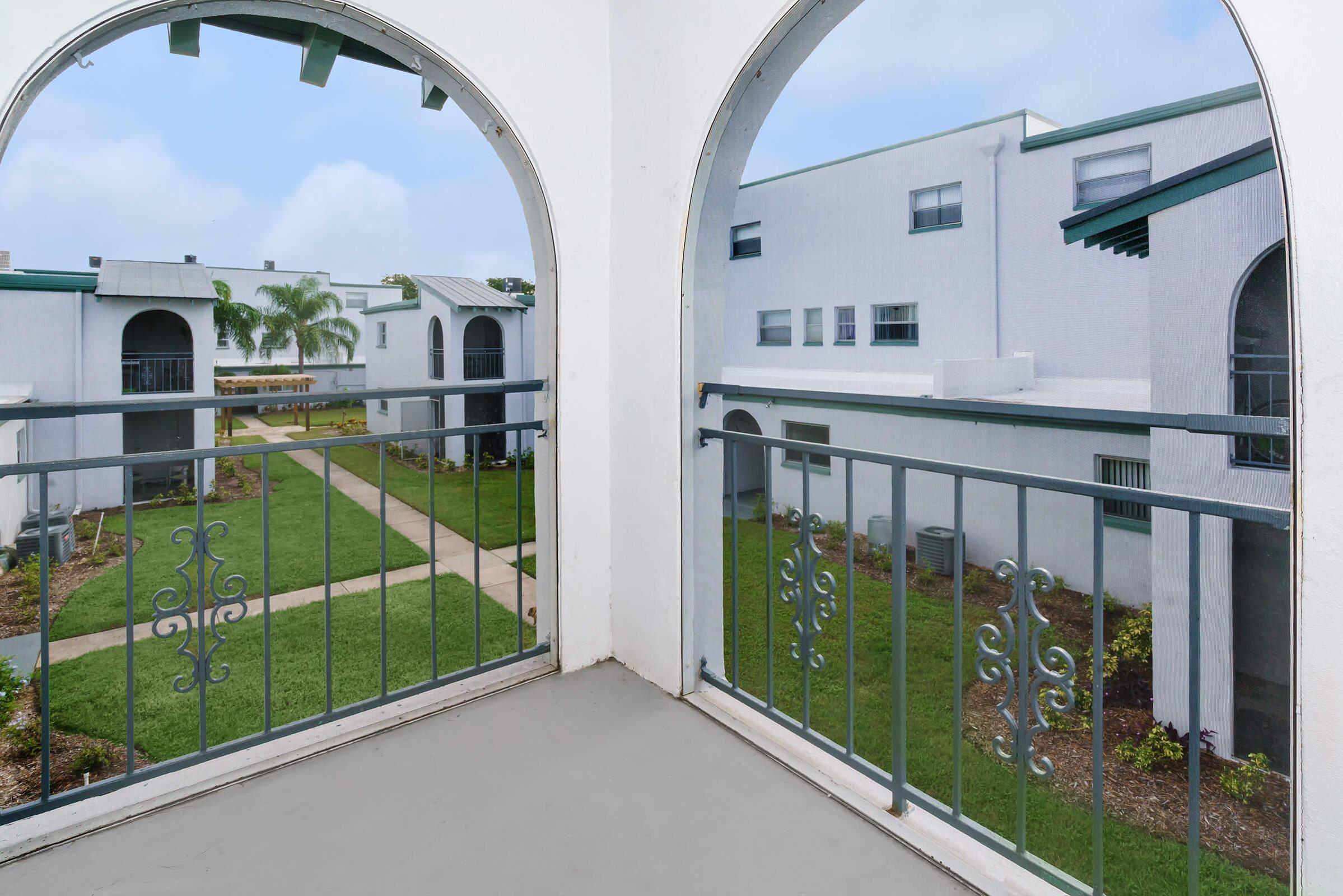 View from a balcony featuring arched openings, overlooking a green courtyard with well-maintained grass and pathways. The buildings in the background are white with green accents, providing a serene and inviting atmosphere.