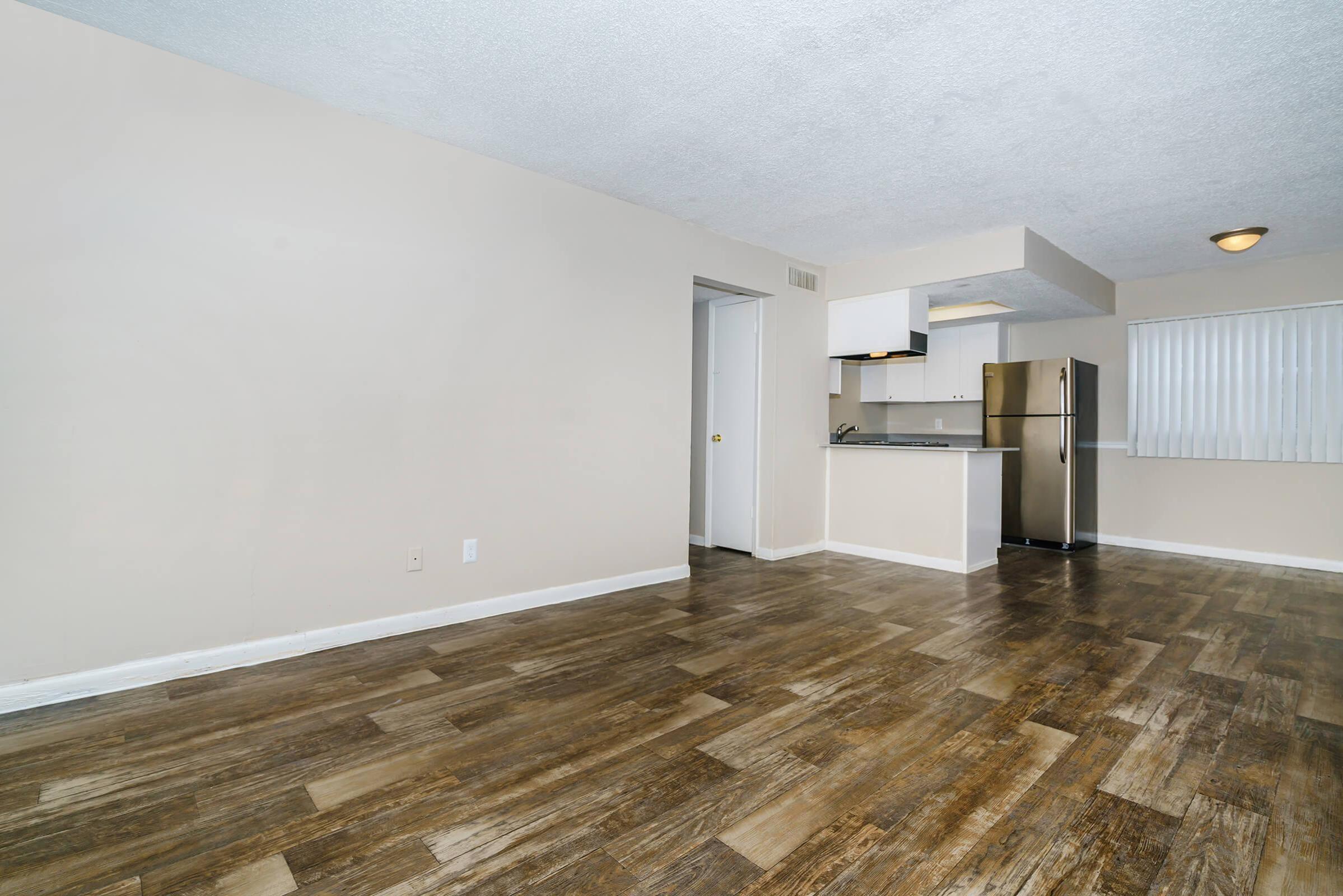 A spacious, minimalist living area featuring hardwood-style flooring, beige walls, and a small kitchen area with stainless steel appliances in the background. There is a white door on the left leading to another room, and a window with blinds allowing natural light to enter.