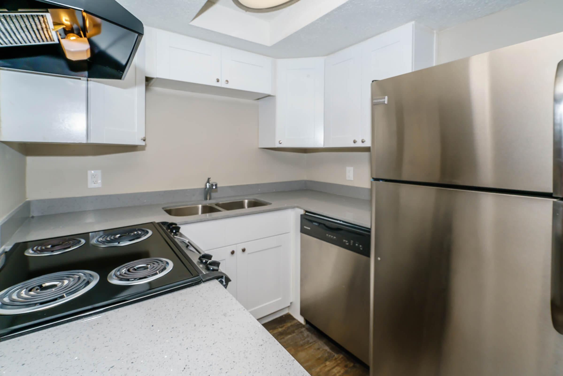 A modern kitchen featuring white cabinets, a stainless steel refrigerator, a dishwasher, and an electric stove with four burners. The countertop is light gray, and there is a double sink with a faucet. The overall design is clean and contemporary.
