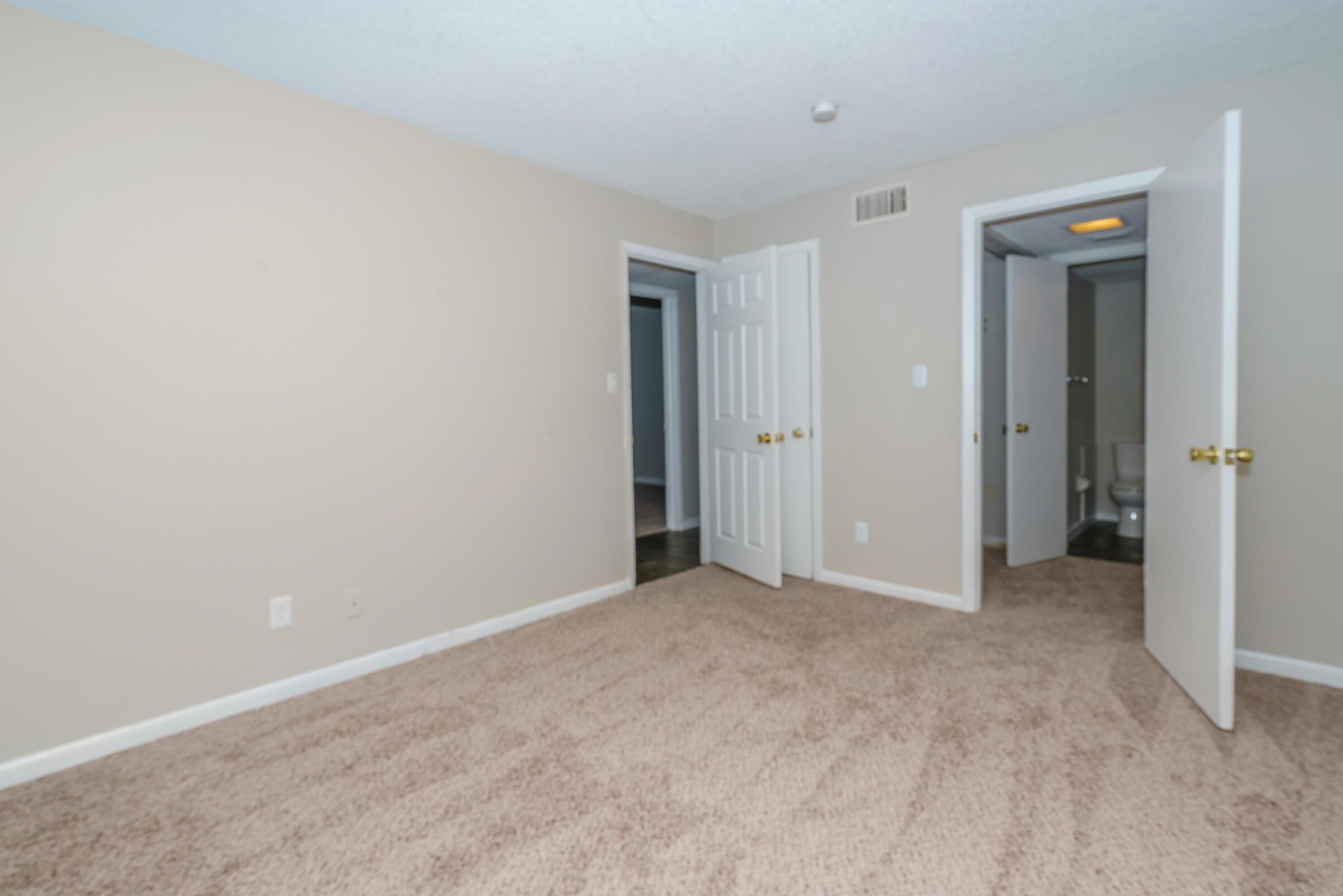A spacious, light-colored bedroom with beige walls and carpet. Two doors lead to adjacent rooms, one partially open revealing a bathroom with a toilet. The room has a clean and neutral aesthetic, suitable for various decor styles. Bright lighting comes from an overhead fixture.
