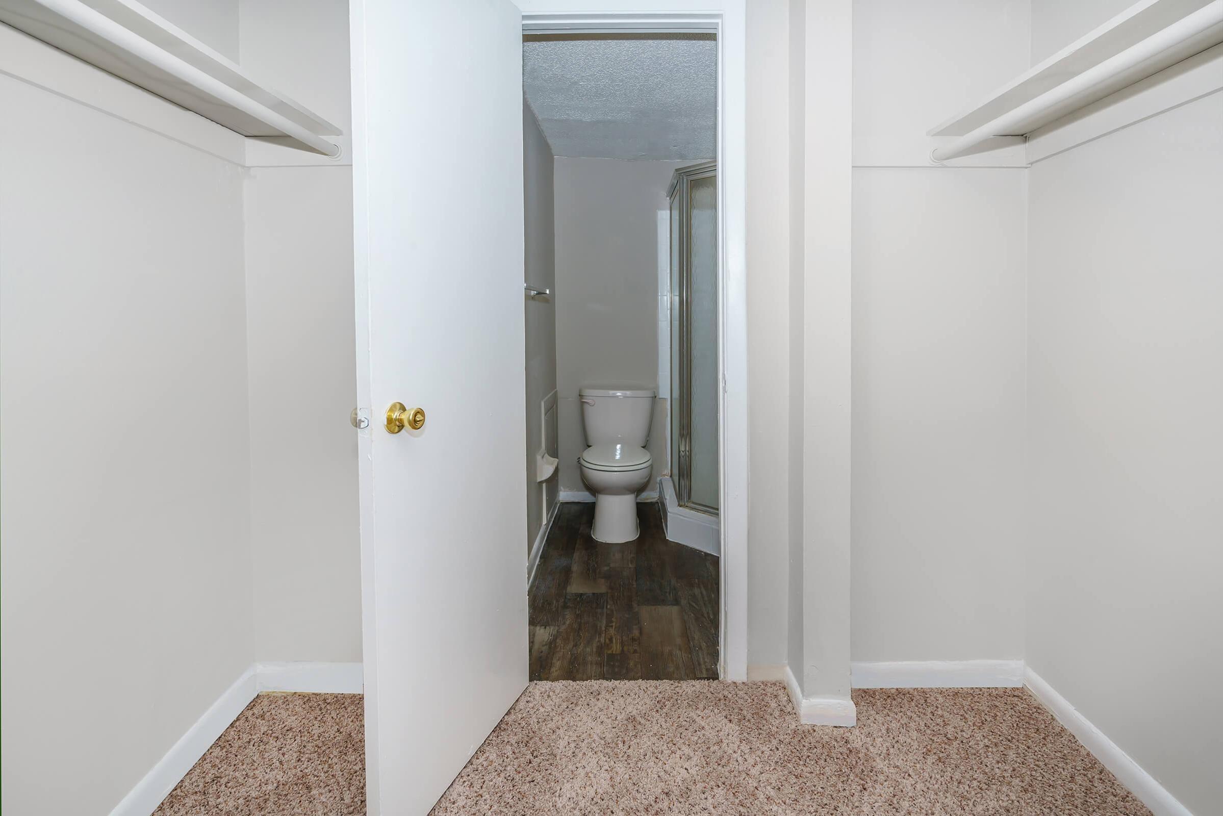 A view of a small, empty room with beige carpet and light gray walls. Open closet spaces on either side lead to a doorway. Beyond the doorway, a bathroom is partially visible, featuring a toilet and a glass shower enclosure. Natural light illuminates the space.