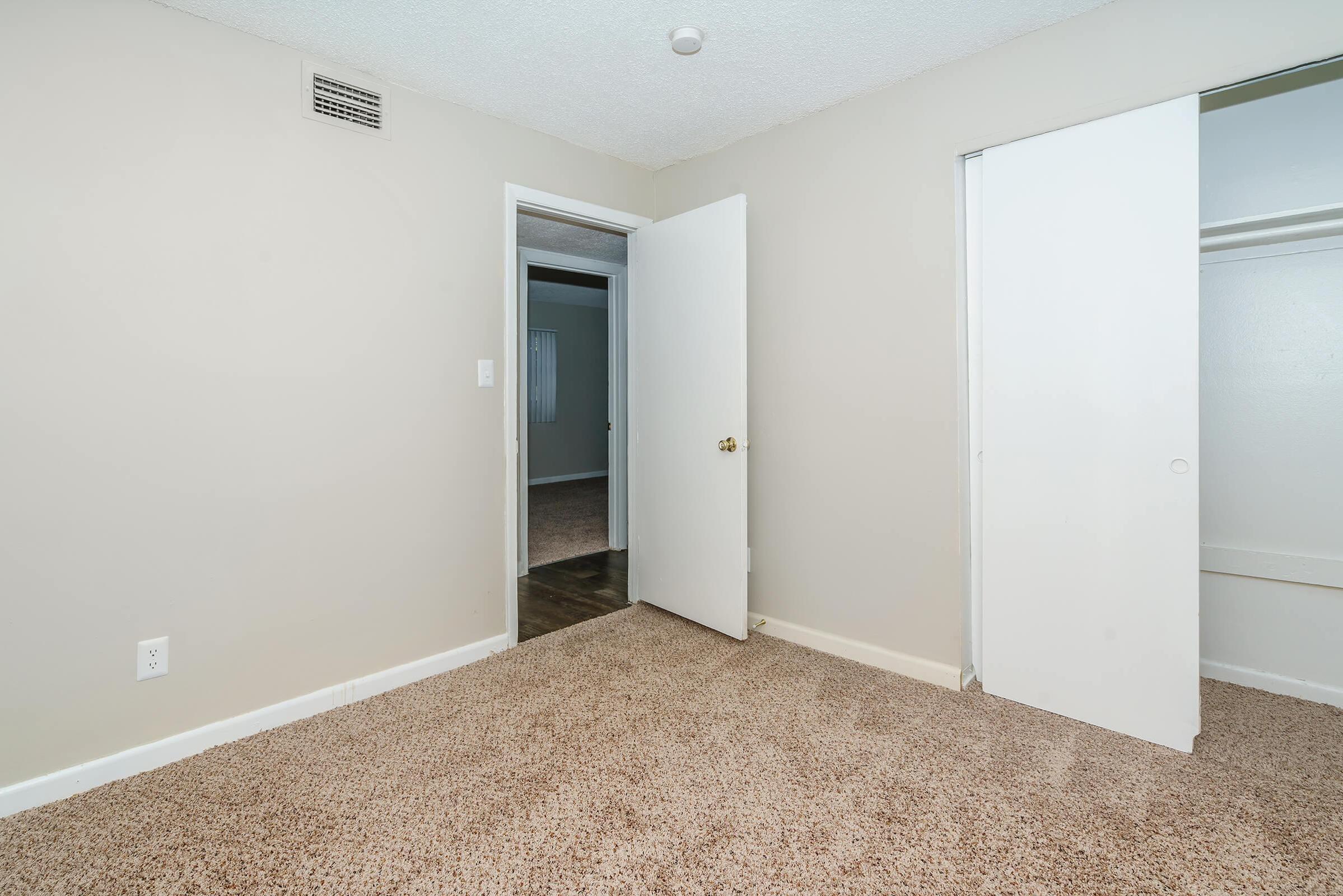 A view of a light-colored room featuring beige carpet, with an open door leading to another area. A closet door is partially opened on the right, revealing a small storage space. The walls are painted a neutral color, creating a simple and airy atmosphere.