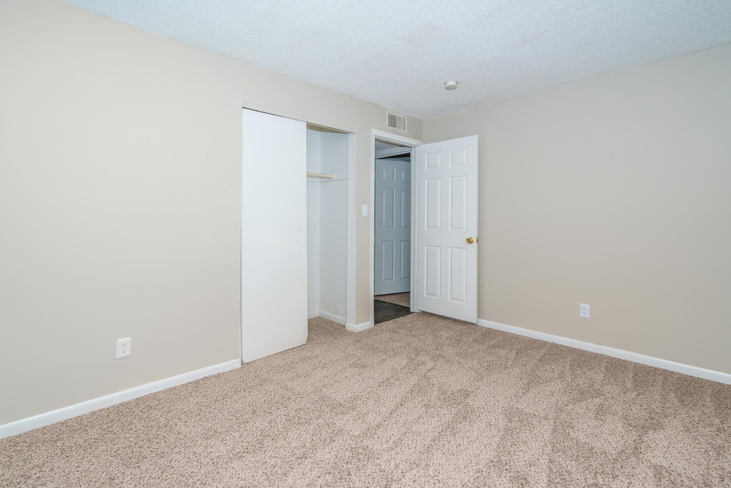 Empty room with beige walls and tan carpet. There are two doors: one leads to a closet and the other to a hallway. The room is well-lit, with a textured ceiling and a simple, uncluttered design, making it suitable for various uses like a bedroom or office space.