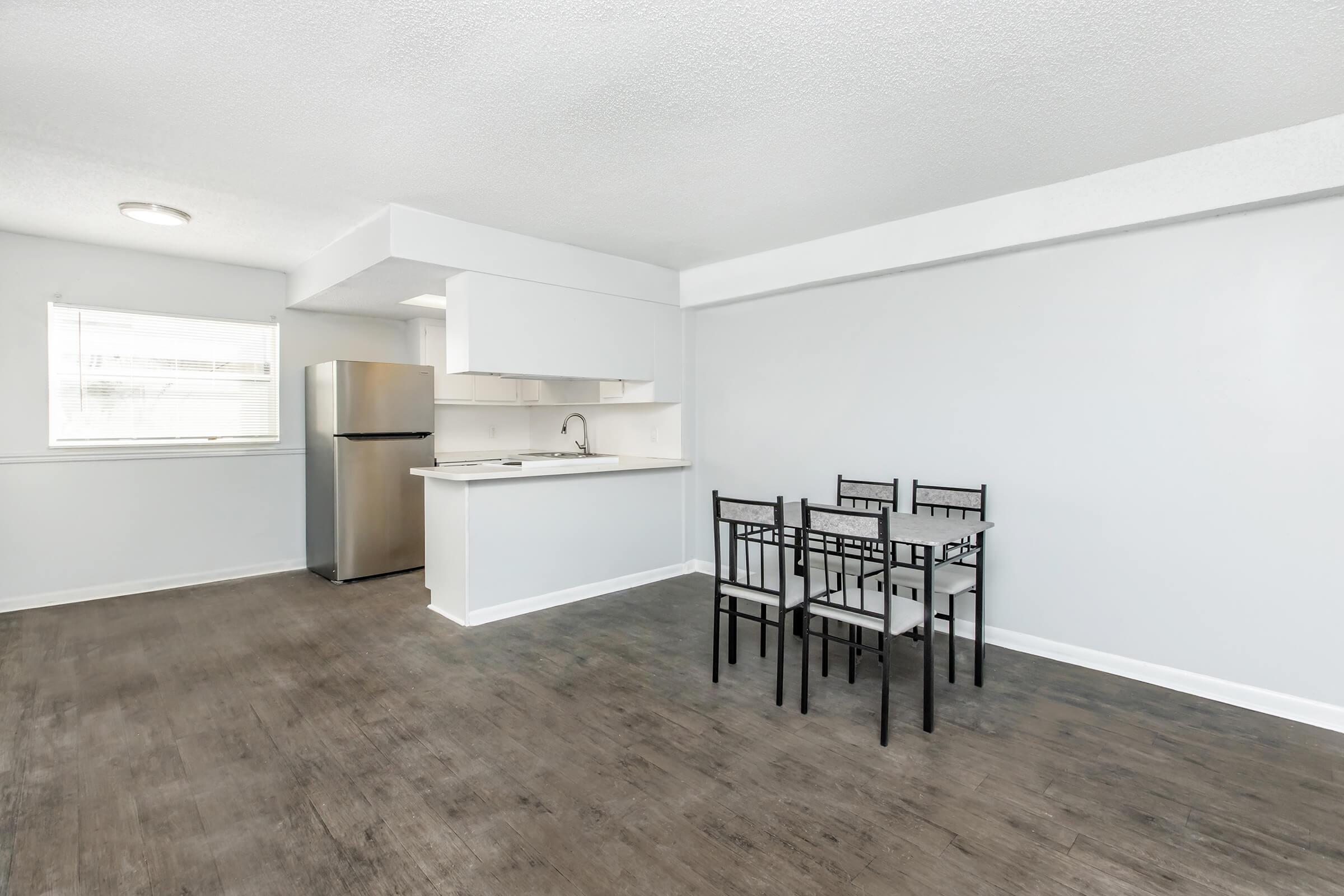 Modern kitchen and dining area in a bright, open-plan space. Features include a stainless steel refrigerator, a simple white kitchen with minimalistic cabinetry, and a small dining table with four black chairs. The floor is a warm brown, and natural light comes through a window.
