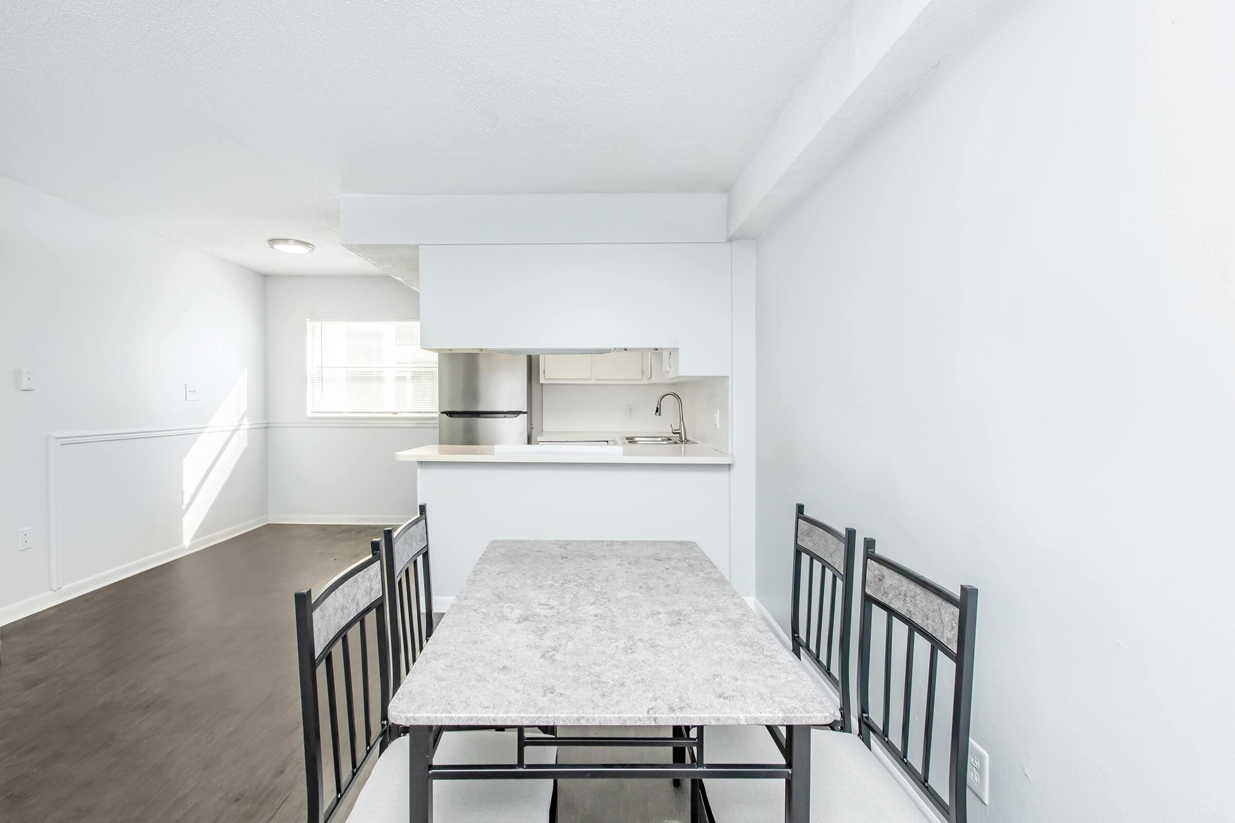 A bright and airy kitchen and dining area featuring a light-colored wall, a small rectangular table with four black chairs, and a kitchen with white cabinetry and stainless steel appliances. Natural light floods the room through a window, creating an inviting atmosphere.
