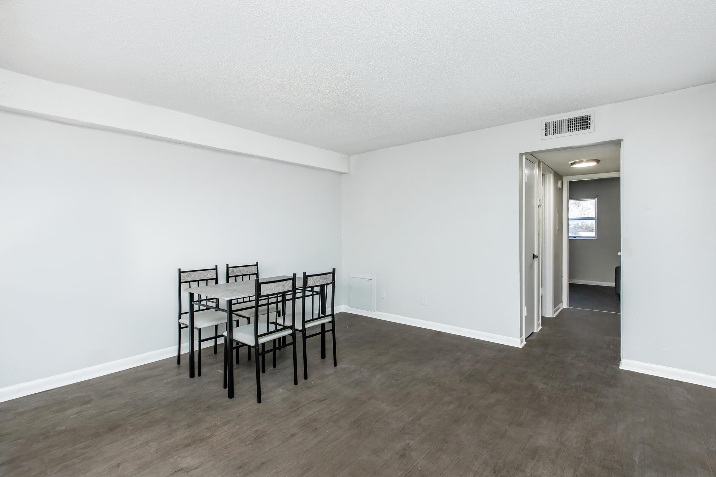 A clean, modern apartment interior featuring a small dining area with a rectangular glass table and four black chairs. The walls are painted white, and the floor has a brown laminate finish. A doorway leads to another room, and there is a ceiling light fixture. Natural light is coming from a window.