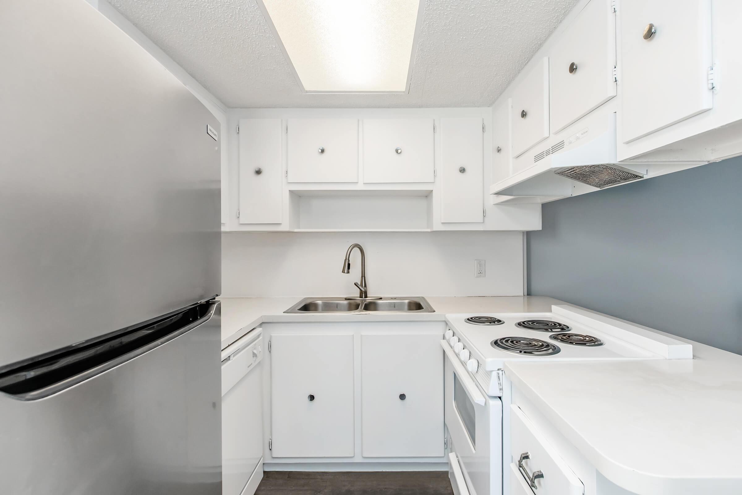 A compact kitchen featuring white cabinetry, a stainless steel refrigerator, a double sink, and a white stove with a vent. The countertop is light-colored, and the kitchen has a modern design with a simple layout and minimal decorations.