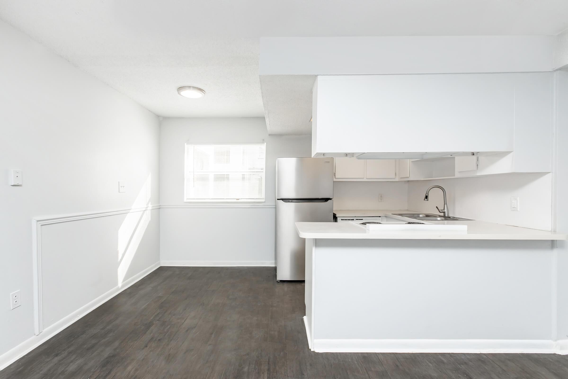 A bright, modern kitchen featuring white cabinets, a stainless steel refrigerator, and a sink with a countertop. The room has light gray walls and a window that lets in natural light, enhancing the spacious and clean atmosphere. The floor is a dark wood finish, providing a nice contrast.