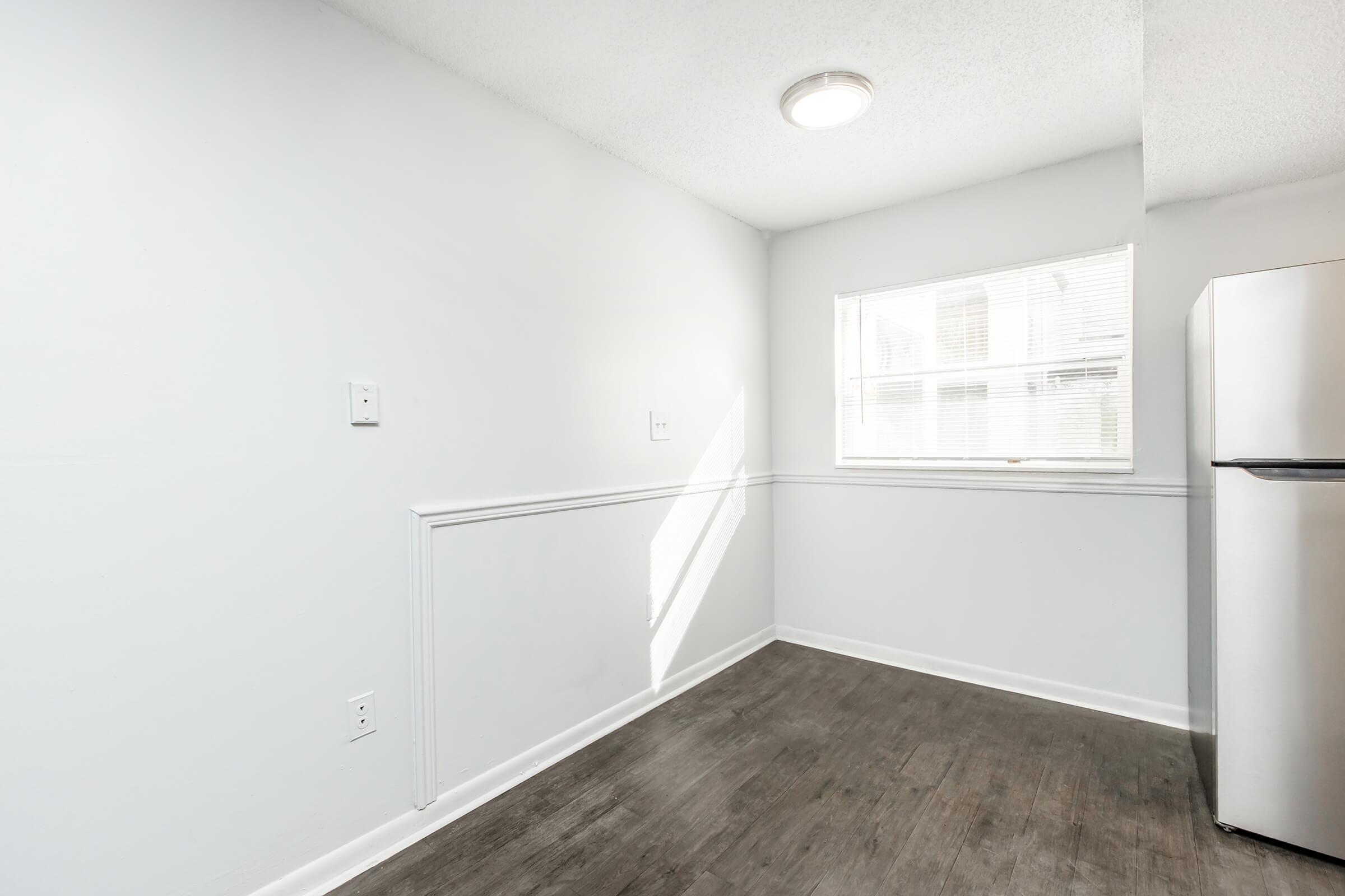 A bright, empty kitchen space with light gray walls and a large window allowing natural light in. The room features a dark wood laminate floor and a stainless steel refrigerator on the right side. The minimalist design includes white trim along the walls and a simple ceiling light fixture.