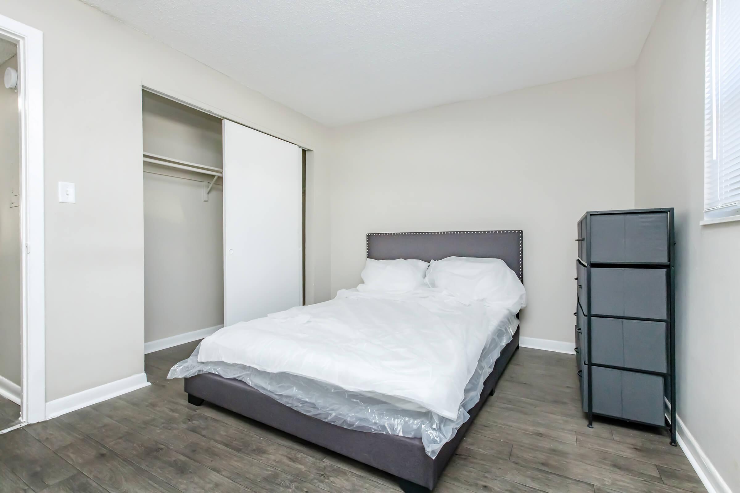 A simple, minimalistic bedroom with a gray upholstered bed covered with white bedding. There is a closet with sliding doors on one side and a small gray storage unit on the other. The room features light-colored walls and a textured floor, creating a clean and tranquil atmosphere.