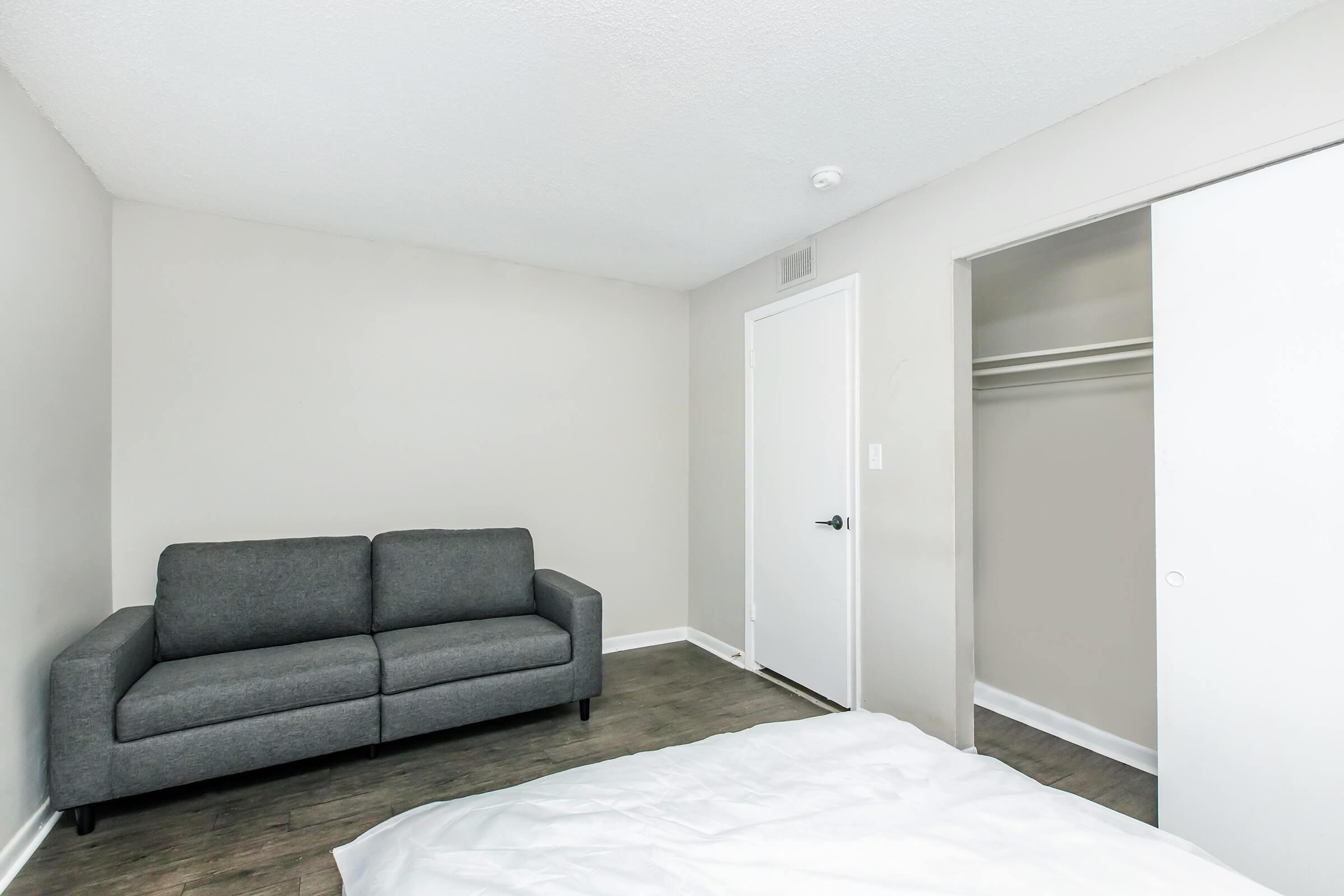 A simple and modern bedroom featuring a gray sofa against a light gray wall, with a closet door visible on the right. The floor has a wood-like finish, and a bed with white bedding is partially visible in the foreground. The overall space is bright and minimalistic.
