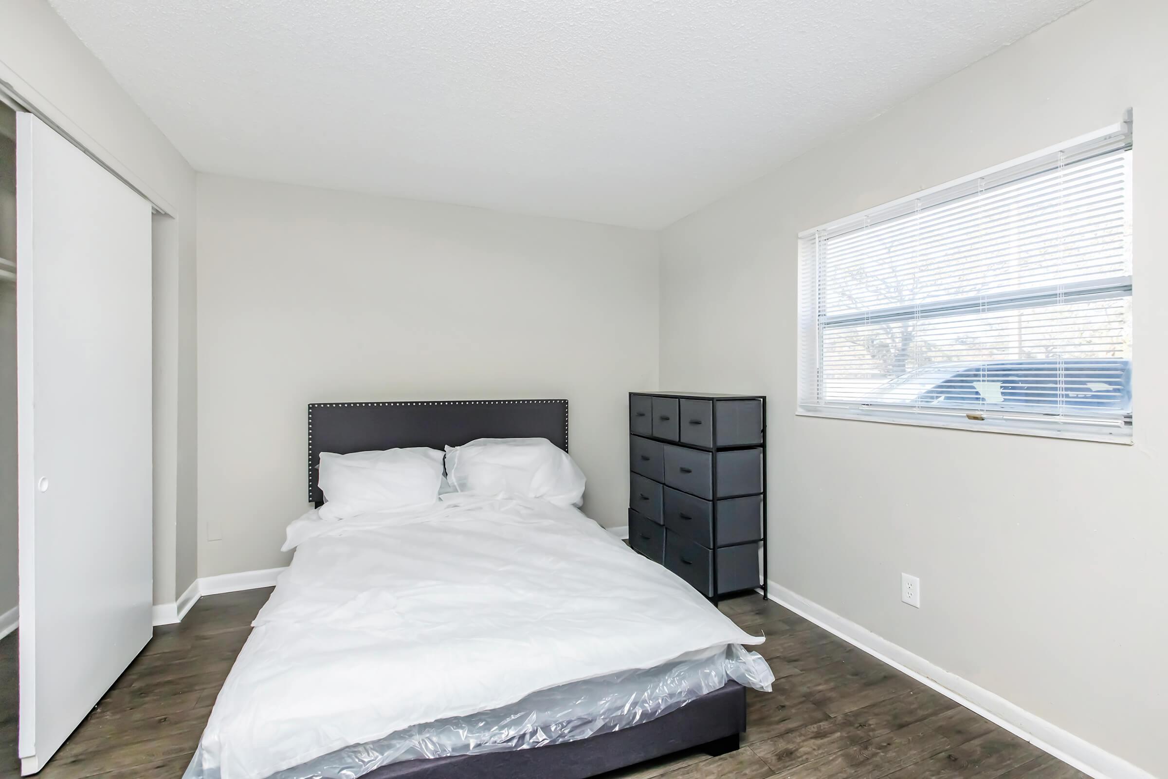 A minimalist bedroom featuring a bed with white bedding, a dark gray headboard, and a dresser with multiple drawers. The walls are painted light gray, and there’s a window with blinds allowing natural light to enter. The floor is a dark wood finish, adding warmth to the space.